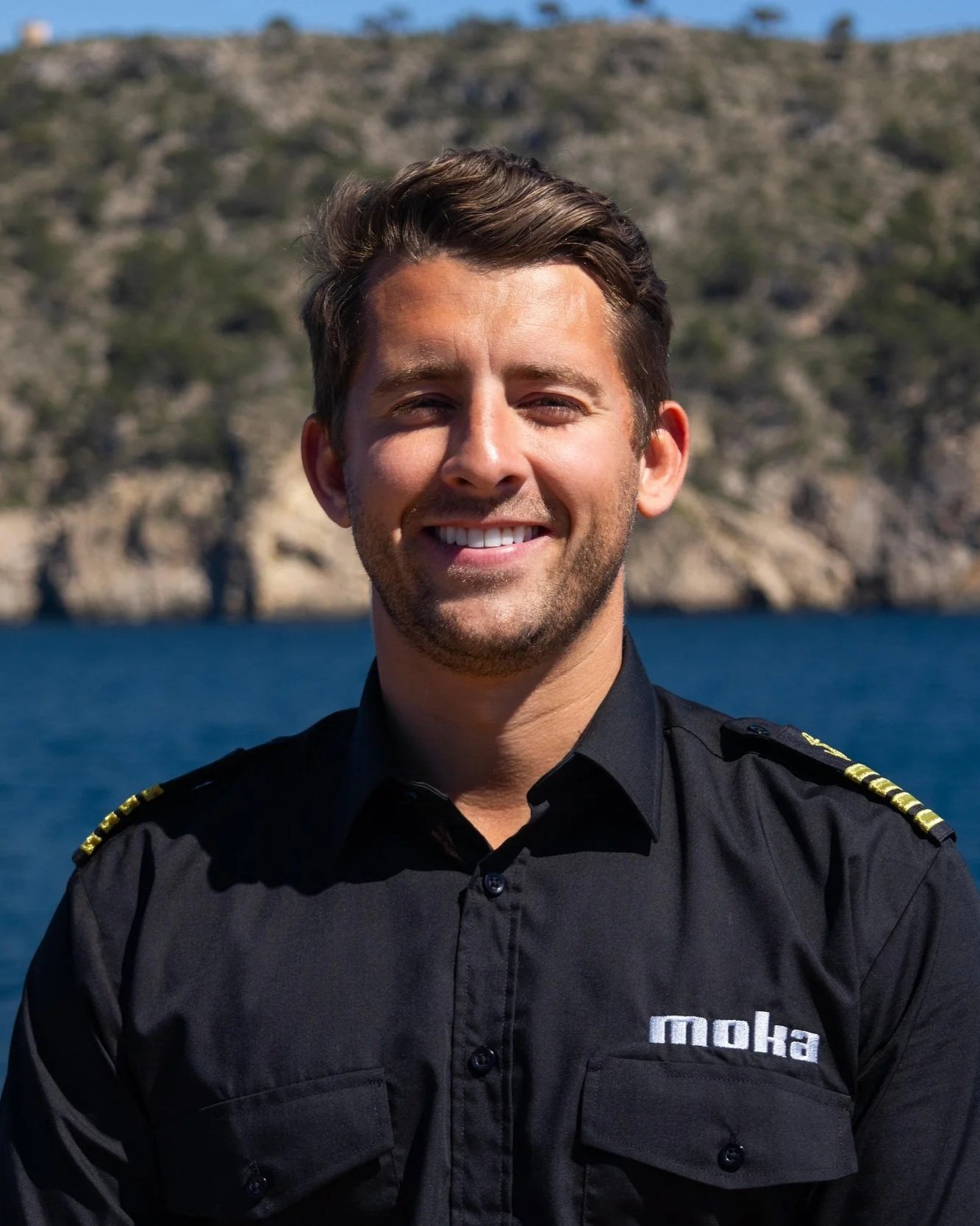 A smiling man in a black uniform with shoulder epaulets, standing outdoors near water with hills in the background.