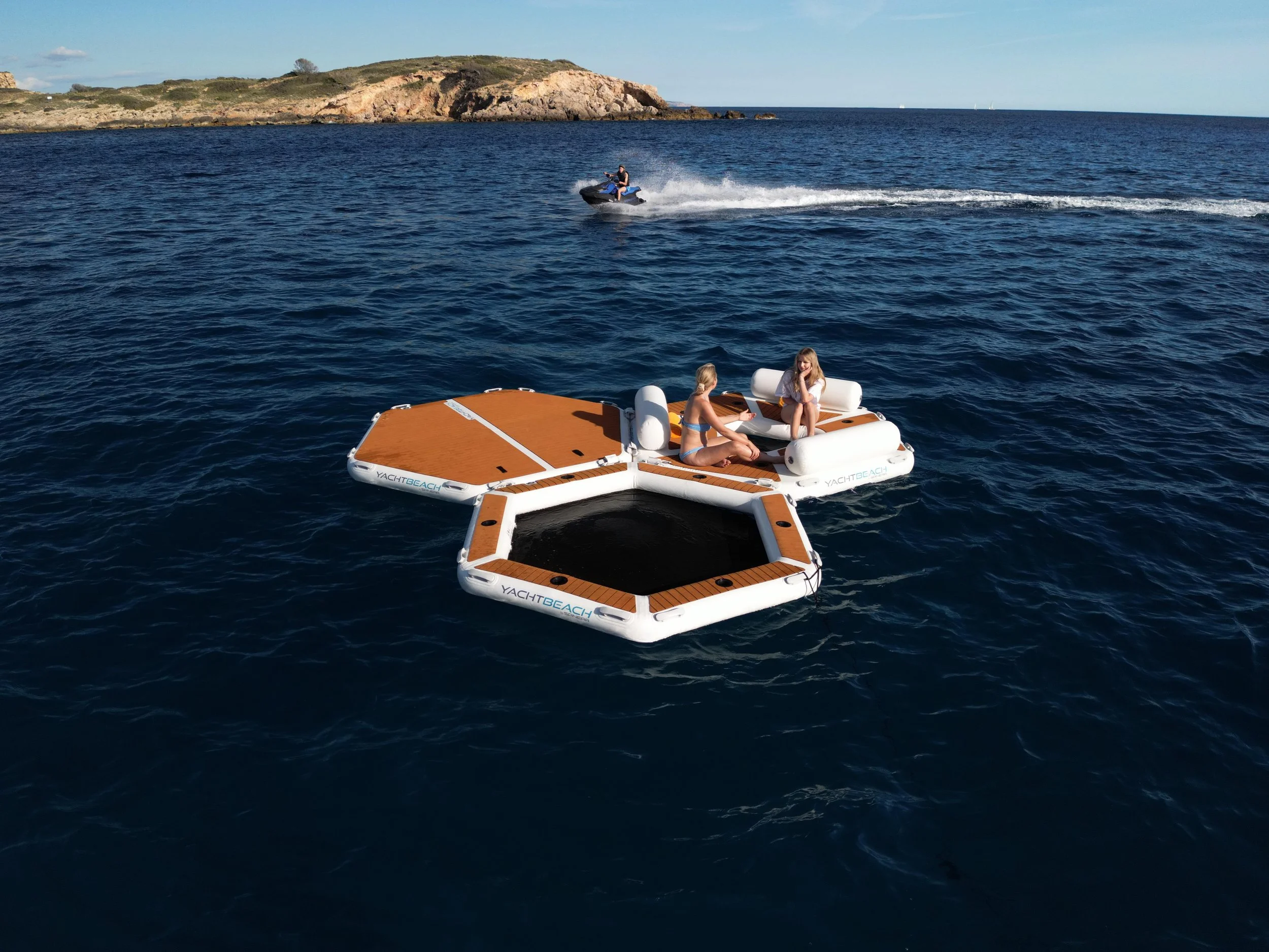 Two women are sitting on a floating platform in the ocean, with a jet ski speeding by in the background and a rocky island on the horizon.