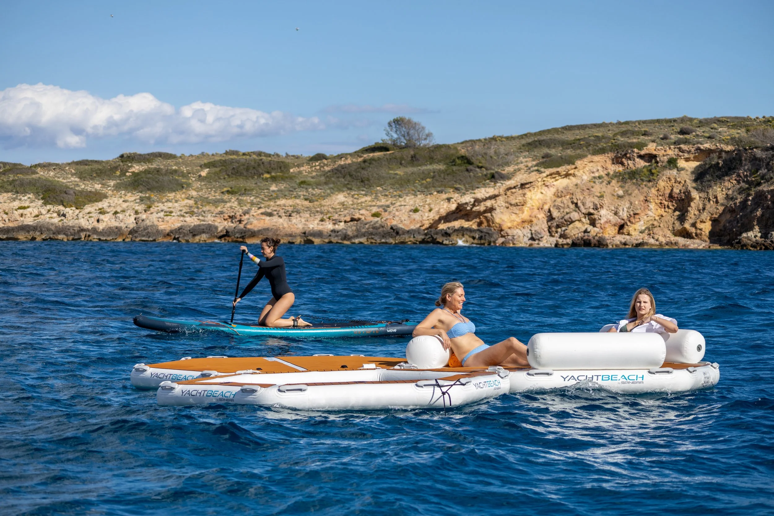 Two women relaxing on paddleboards and a woman kneeling on a paddleboard, all on calm water near rocky shoreline with hills and blue sky.