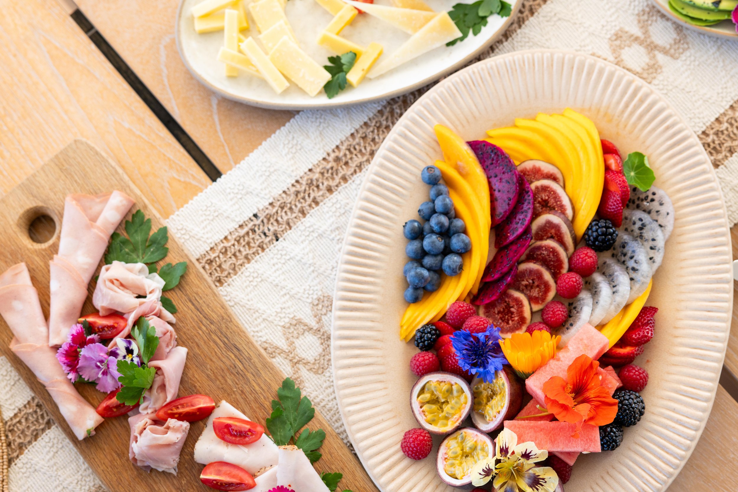 Colorful fruit platter with blueberries, blackberries, raspberries, strawberries, watermelon, dragon fruit, and passion fruit, served on a white oval dish. There is also a cheese and cheese, and vegetable platter with cherry tomatoes, herbs, and rolled deli meats on a wooden board.