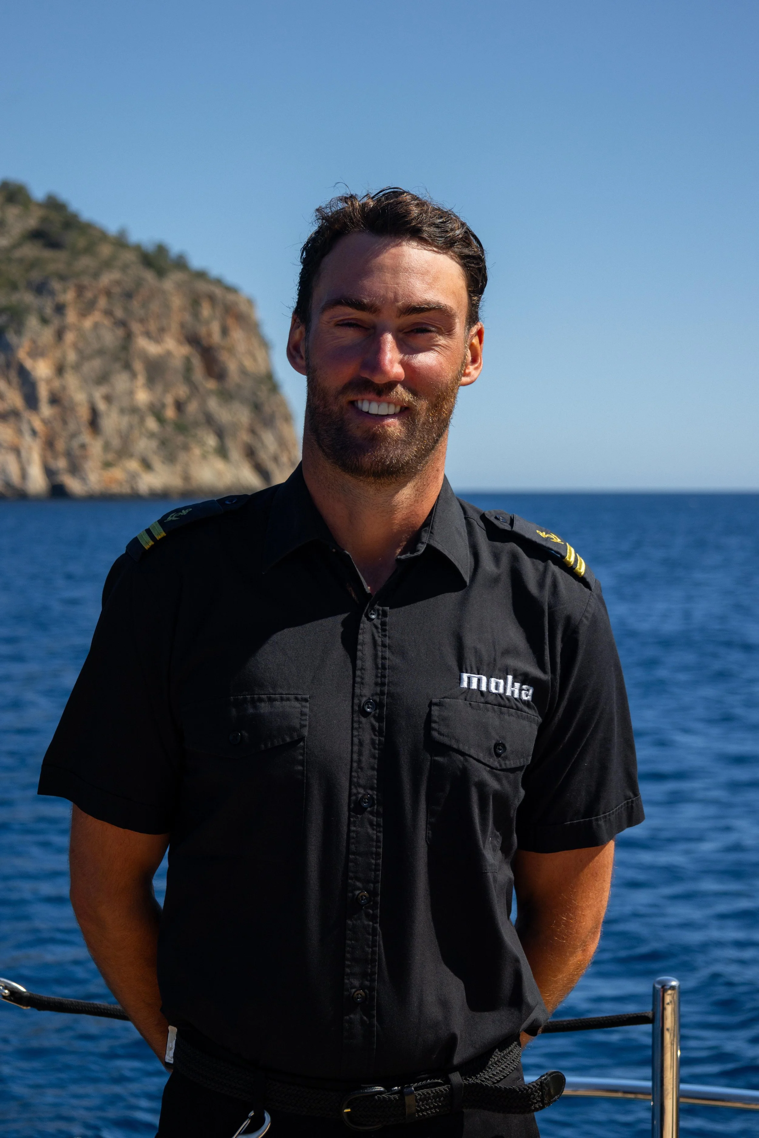A man smiling on a boat, wearing a black uniform with 'moha' embroidered on the chest, with a rocky coastline and blue water in the background beneath a clear blue sky.