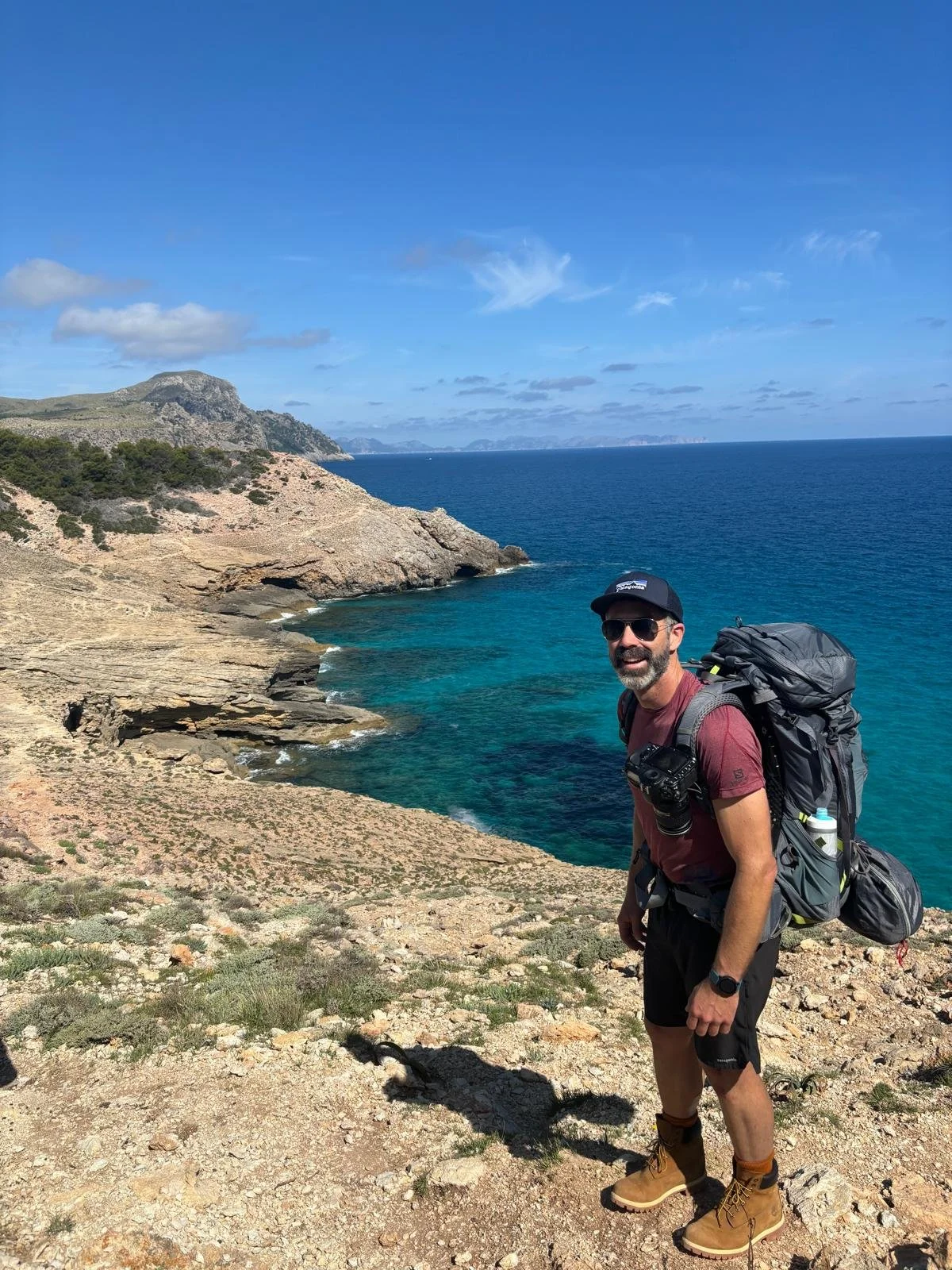 A man with a beard, sunglasses, and a baseball cap, carrying a large backpack, standing on a rocky coastline with clear blue water and a partly cloudy sky.
