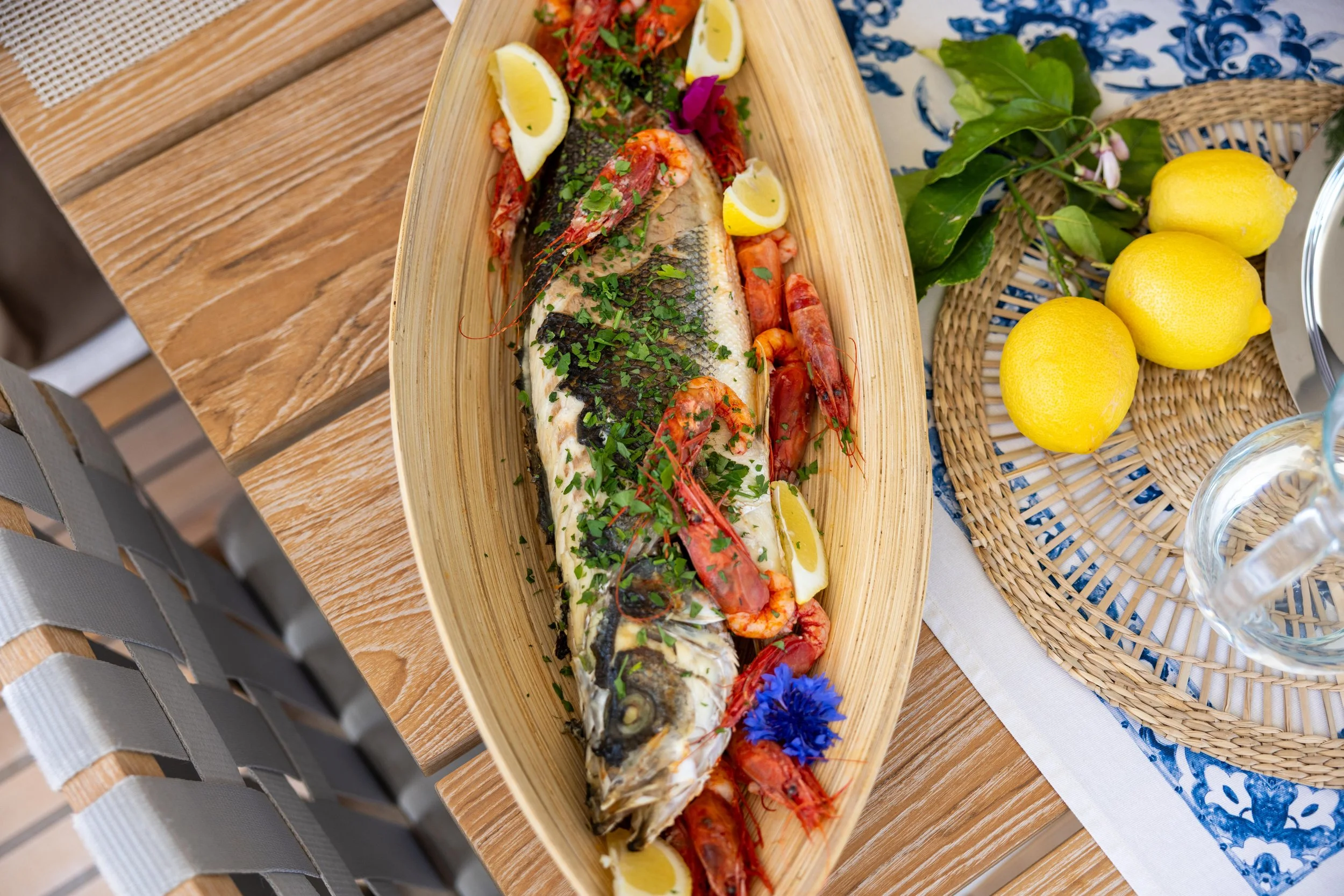 A serving of grilled fish with shrimp and lemon wedges garnished with herbs on a wooden platter, next to a basket of lemons, greenery, and a glass of water on a table.