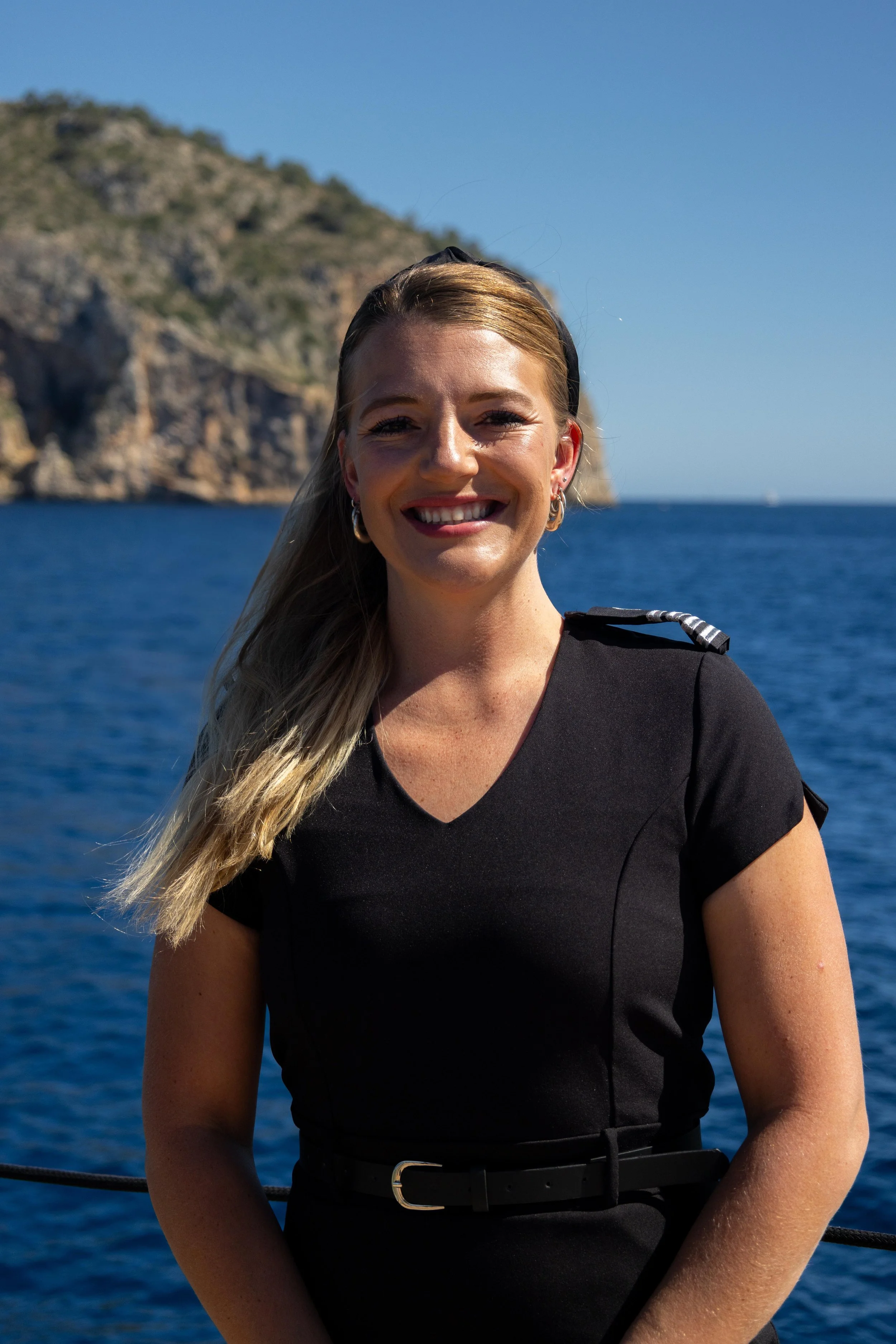 A smiling woman with blonde hair, wearing a black uniform with a shoulder epaulet, standing outdoors near a body of water with cliffs in the background.