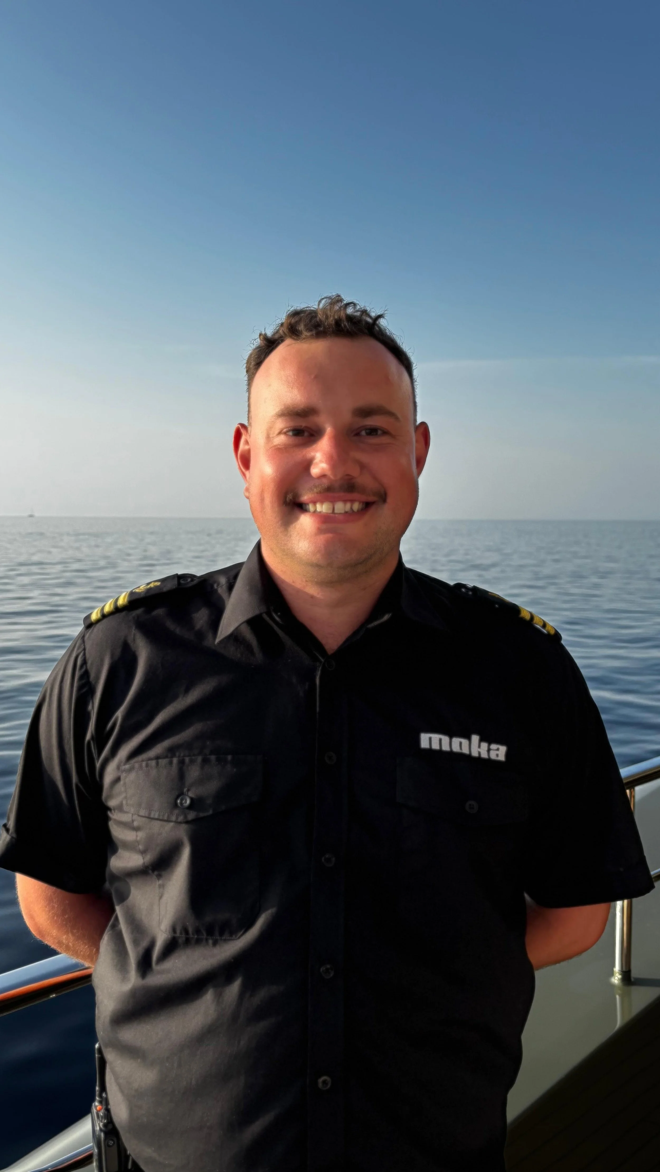 A smiling man in a black uniform with yellow epaulets, standing on a boat with the ocean and sky in the background.
