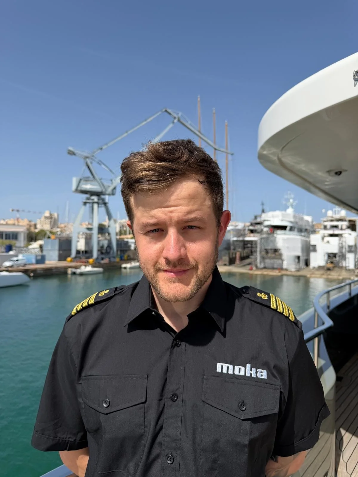 A young man with short brown hair, a beard, and a slight smile in a black uniform with gold shoulder epaulets, standing on a boat near a marina with yachts, a crane, and buildings in the background under a clear blue sky.