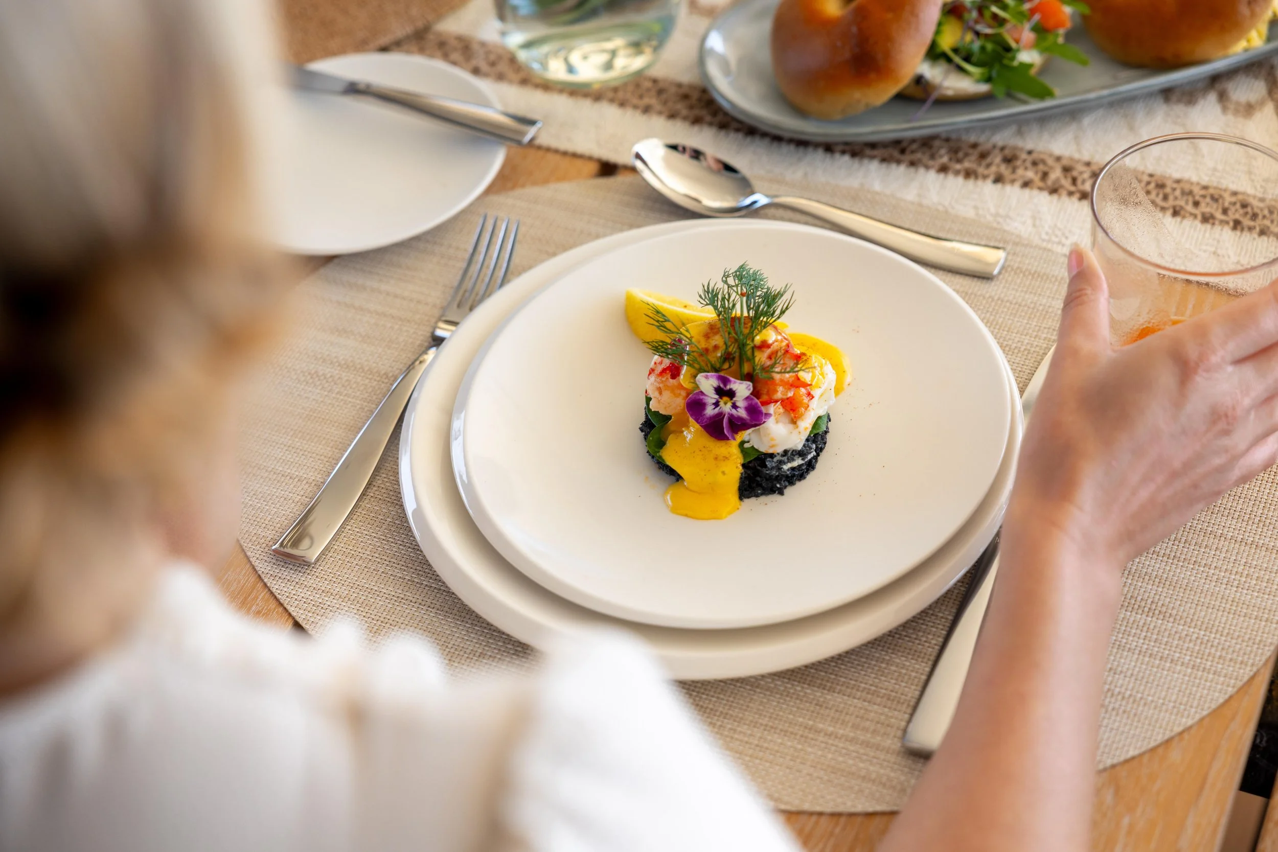 A woman at a formal dining table with a plate of gourmet food, garnished with herbs and edible flowers, and a glass of beverage.