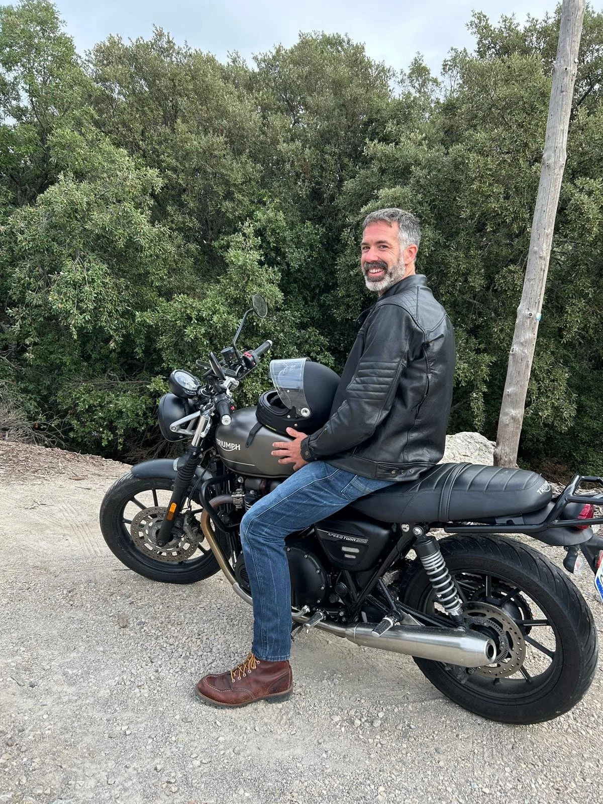 A smiling man in a black leather jacket and blue jeans sitting on a black Triumph motorcycle on a gravel road with green bushes and a wooden utility pole in the background.
