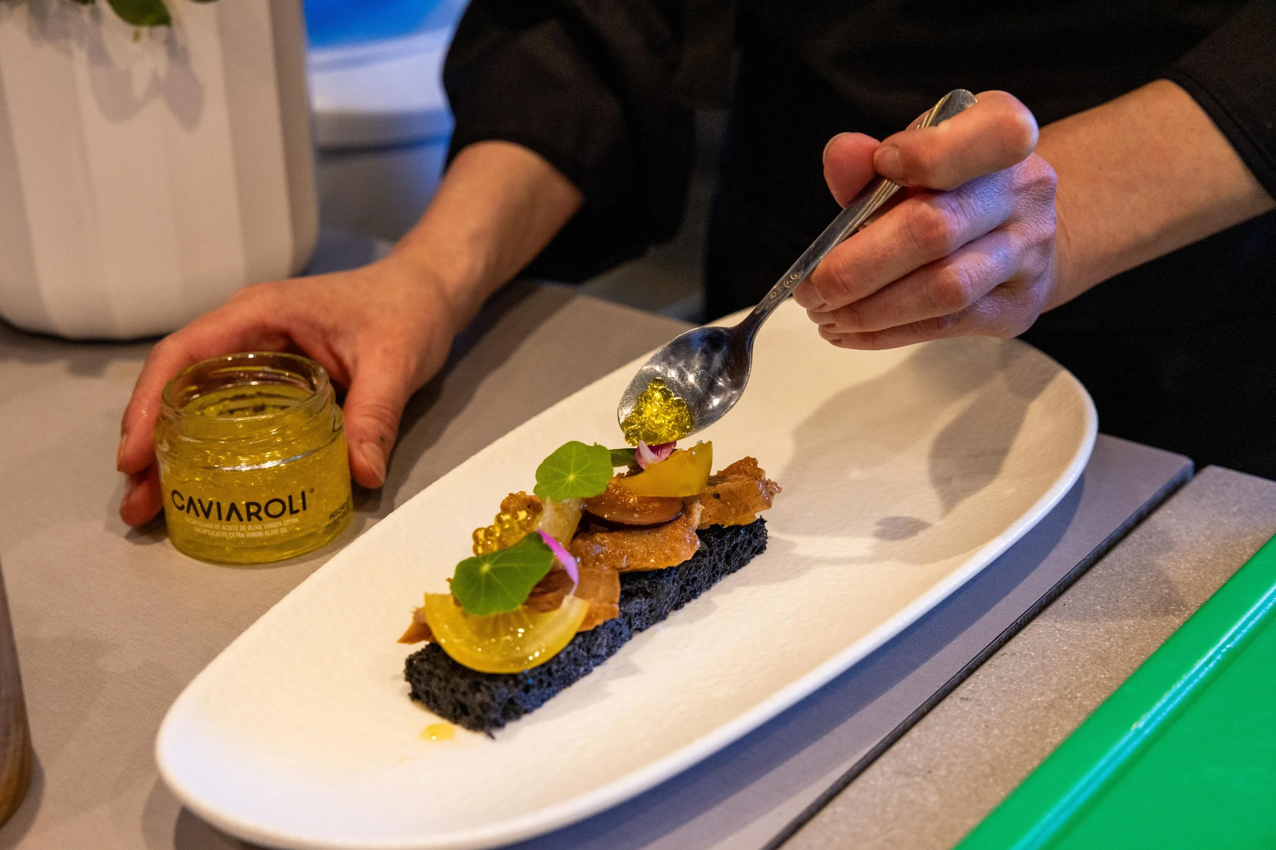 Person drizzling yellow caviar onto a gourmet appetizer with black bread, yellow tomato, microgreens, and edible flowers on a white oval plate.