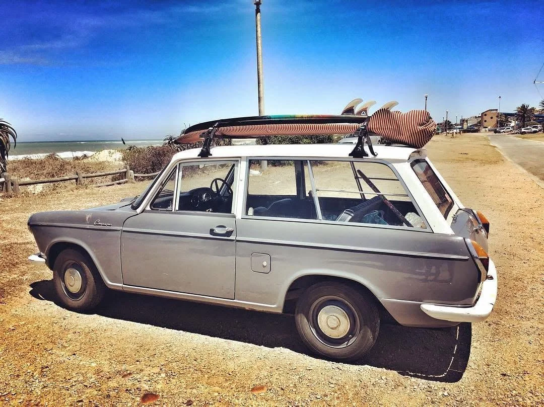 A vintage silver station wagon parked on a dirt road near the beach, with a surfboard on the roof.