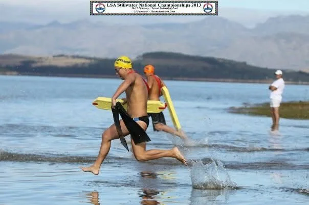 Young male swimmer running out of water with a yellow kickboard, wearing a yellow swim cap and black swimming trunks, during a competition at a lake or river.