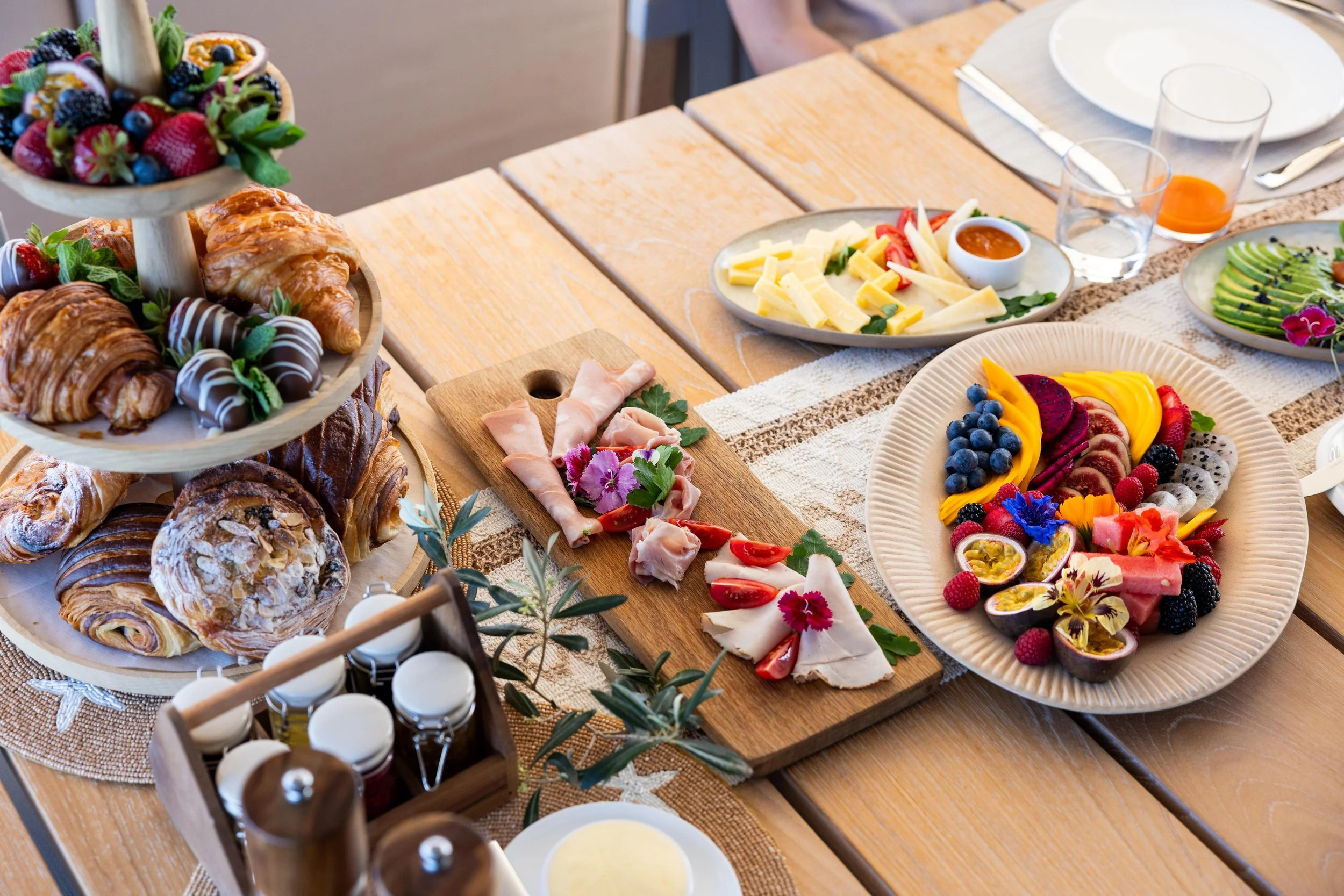 A breakfast table with a tiered stand of croissants, Danish pastries, and muffins, a platter of sliced meats and cheeses, a colorful fruit platter, and various plates with cheese and avocado slices.