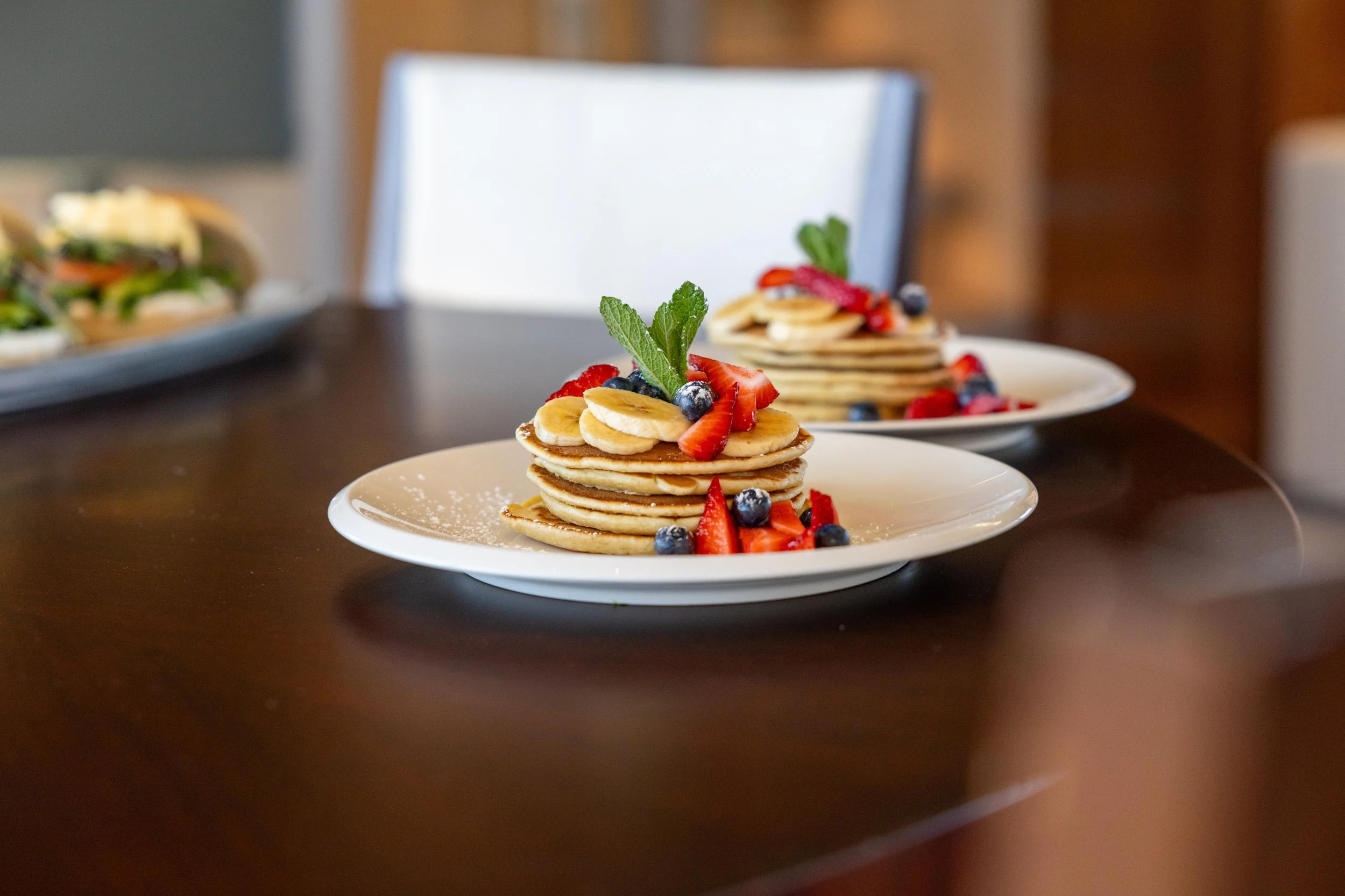 Piles of pancakes topped with sliced bananas, strawberries, blueberries, and fresh mint on a white plate.