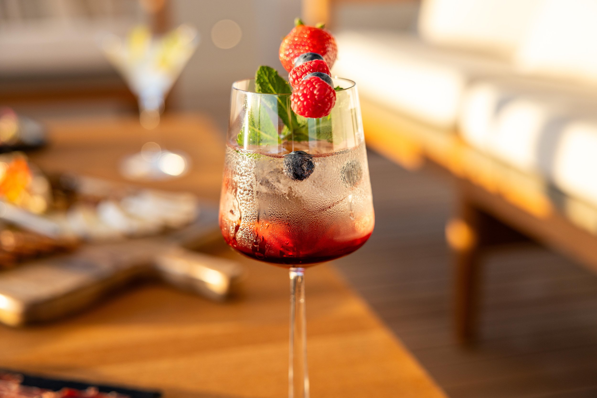 A tall glass with a pinkish red cocktail garnished with strawberries, blueberries, mint leaves, and ice cubes, on a wooden table.