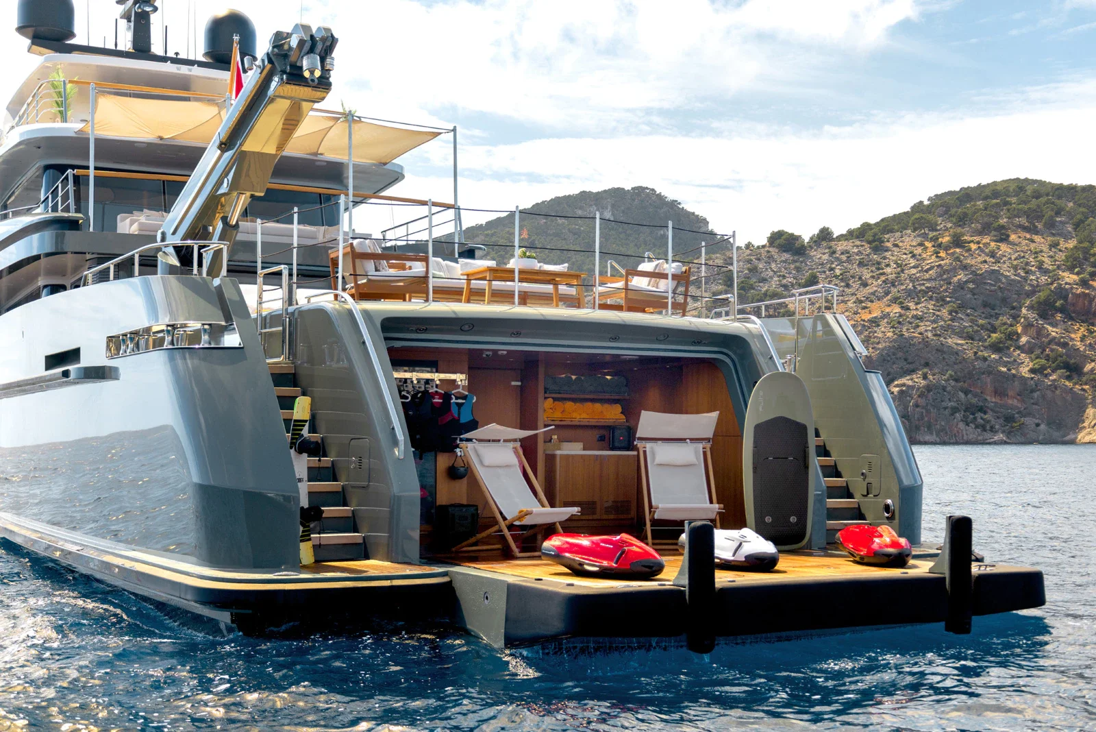 A luxurious yacht on the water with a scenic mountain background, featuring an open deck area with lounge chairs, small remote-controlled boats, and swimming gear.