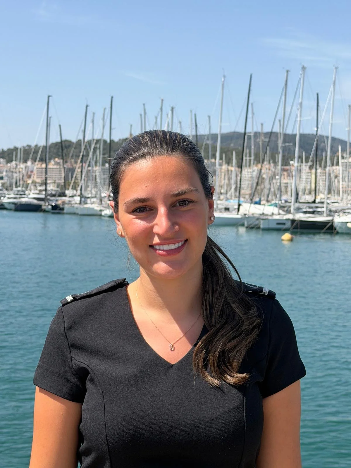 A young woman with long dark hair, smiling, wearing a black top, standing in front of a marina with many sailboats and a hillside in the background.