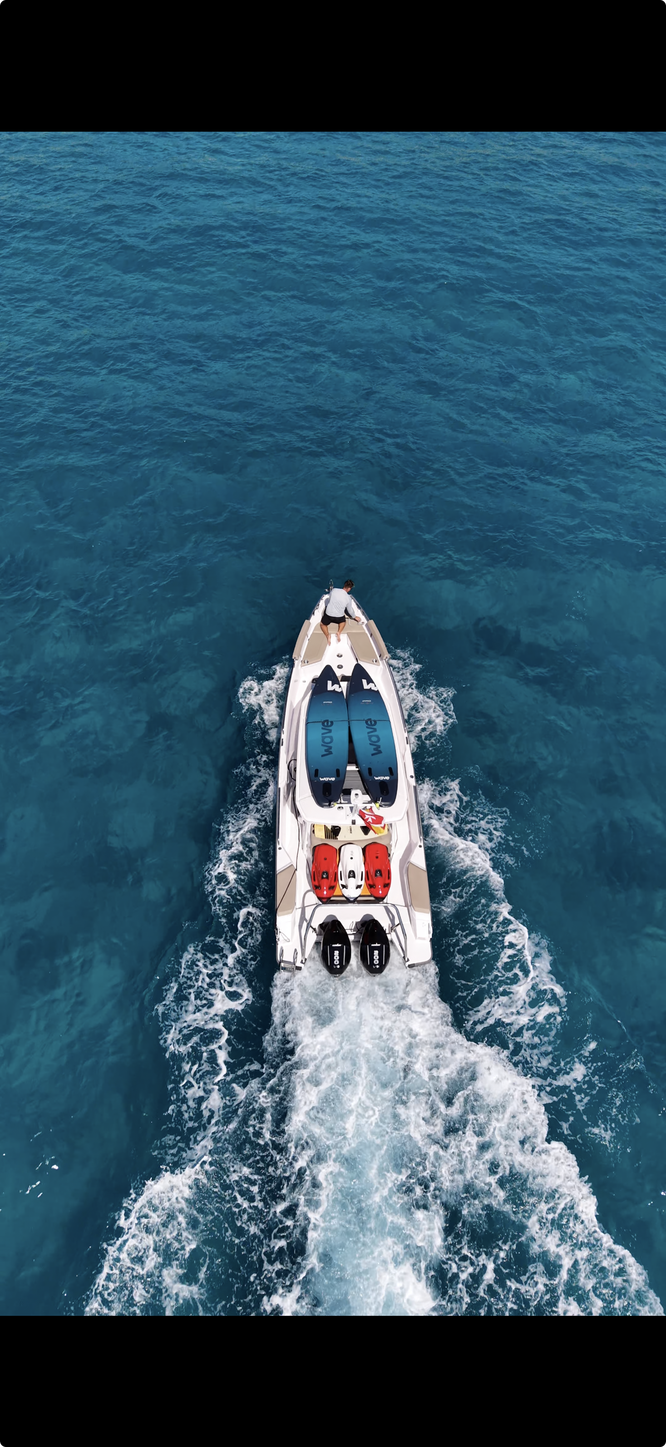 A person standing at the bow of a speedboat on the open ocean, with watercraft equipment including water skis and jet skis secured on the boat.
