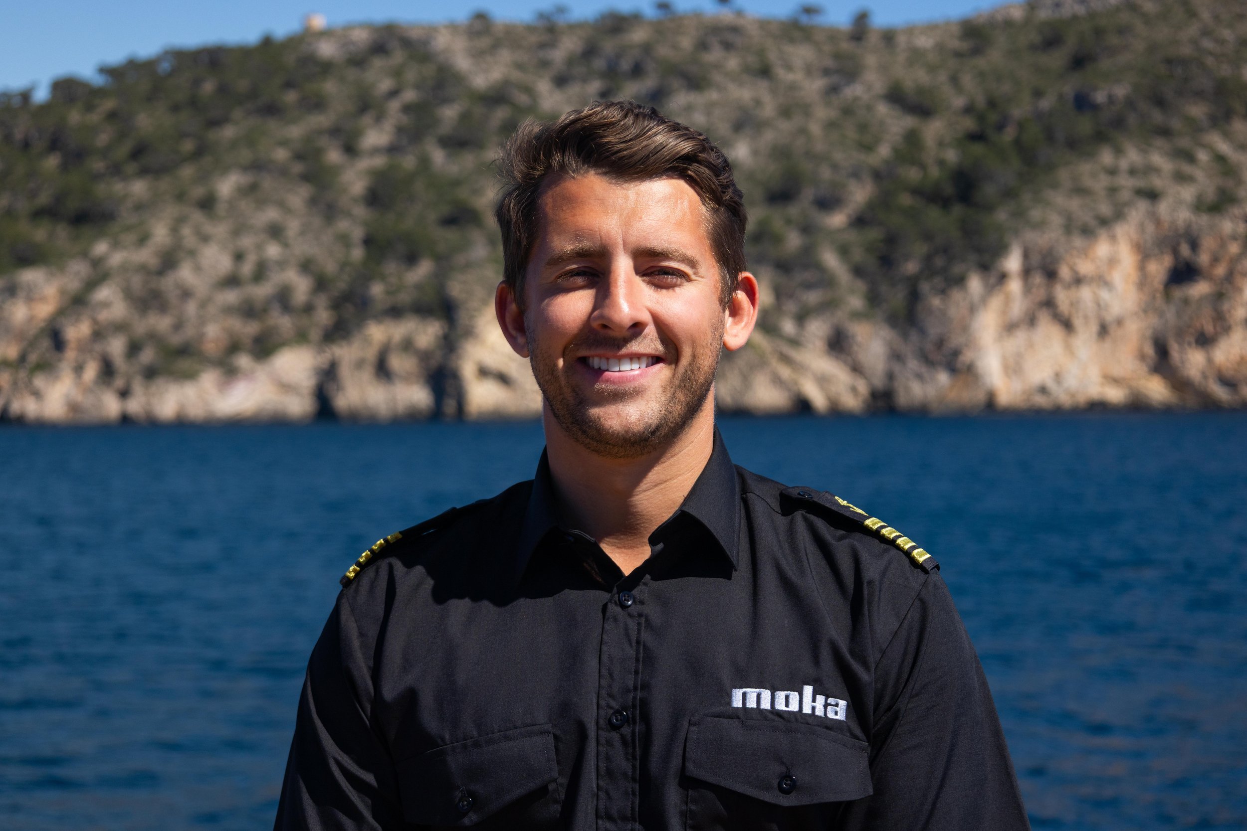 A smiling man in a black uniform standing outdoors near a body of water with rocky hills in the background.