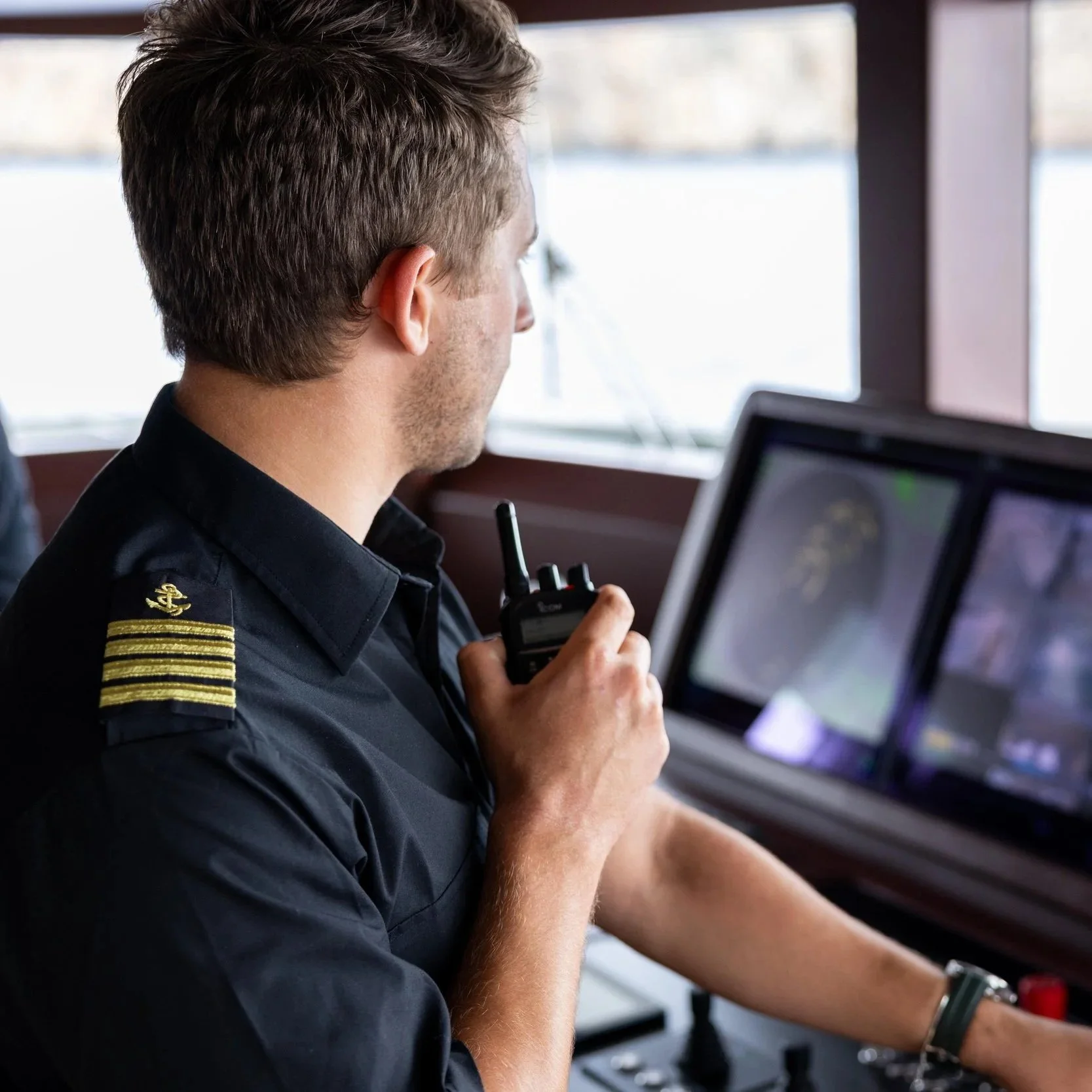 A man in a navy-blue uniform with gold stripes on the shoulder, sitting in front of a computer monitor, holding a radio device, with a window in the background.