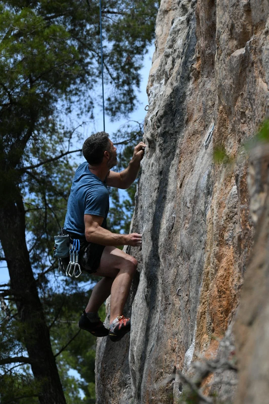 A man rock climbing on an outdoor vertical rock face, wearing a blue shirt, shorts, and climbing shoes, with climbing gear attached to his waist, surrounded by trees and a blue sky.