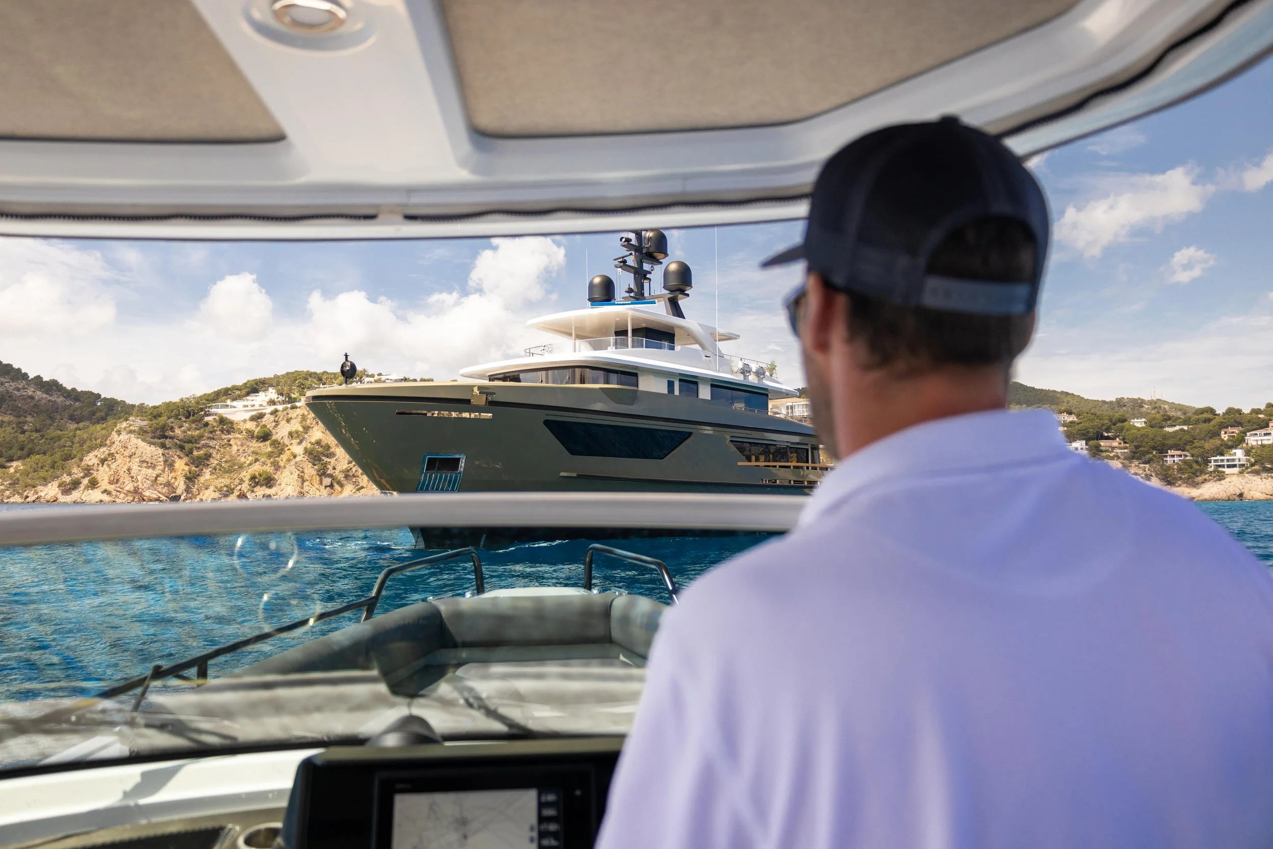 A man wearing a black cap and white shirt looking at a large yacht sailing in the water against a hillside with houses, under a partly cloudy sky.