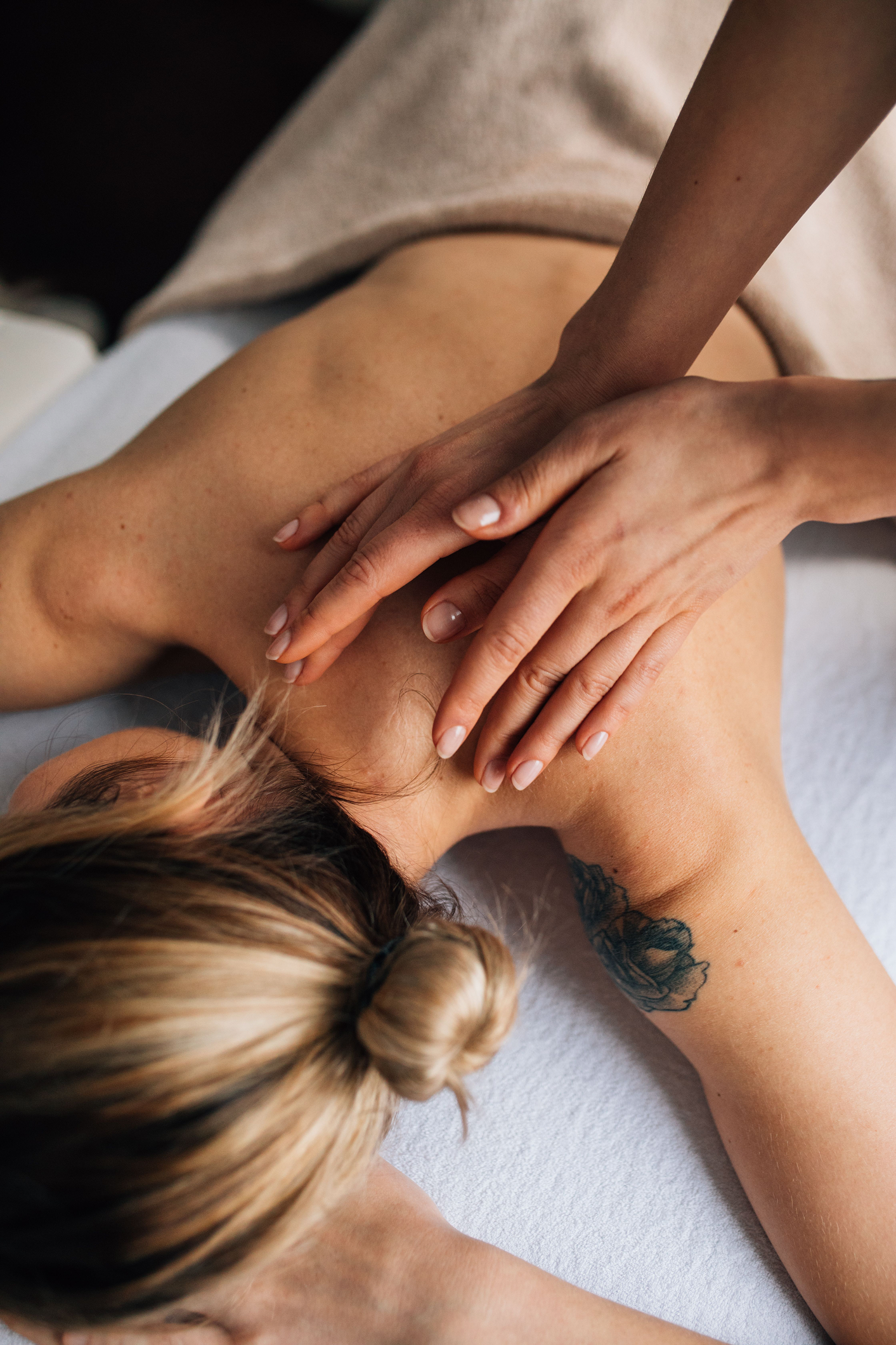 A person receiving a massage, lying on their stomach with their head turned to the side. The person's hand is resting on their back, and they have a tattoo of flowers on their arm. Their hair is tied up in a bun.
