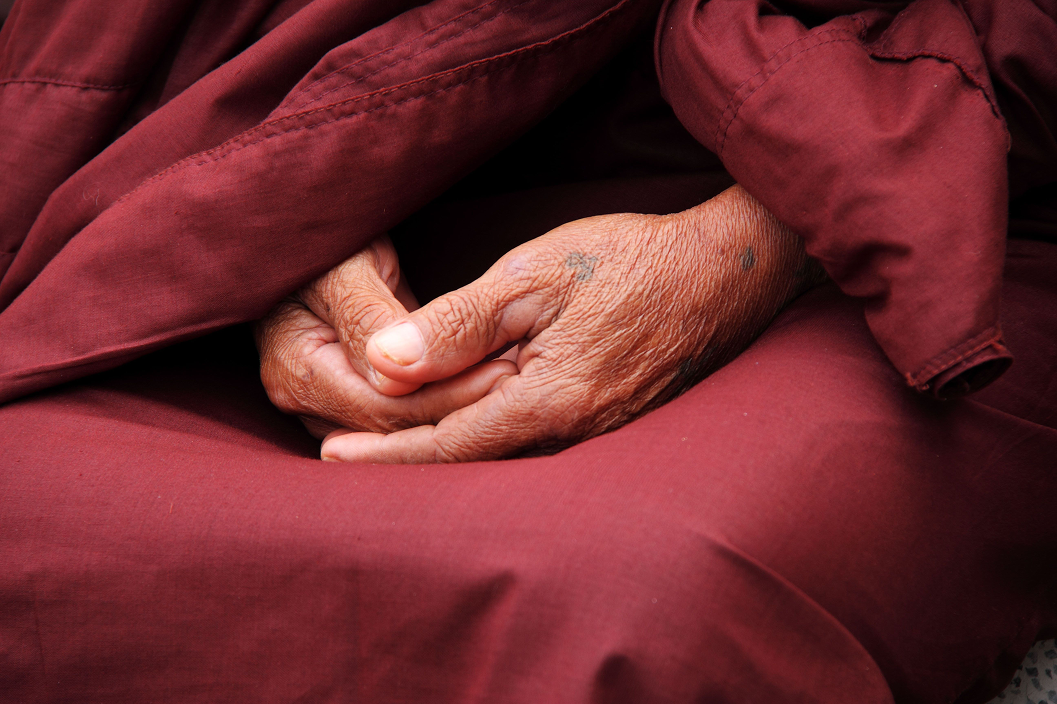 Close-up of an elderly person's hands resting in their lap, with visible age spots and wrinkles, wearing reddish-brown clothing.