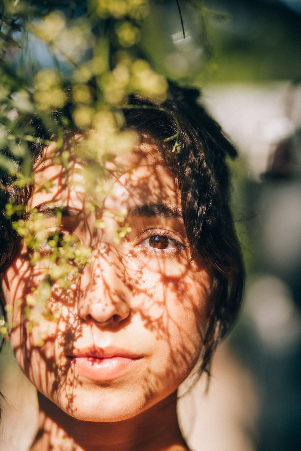 A young person with short dark hair and brown eyes, partially covered by leafy branches casting shadows on their face, outdoors in sunlight.
