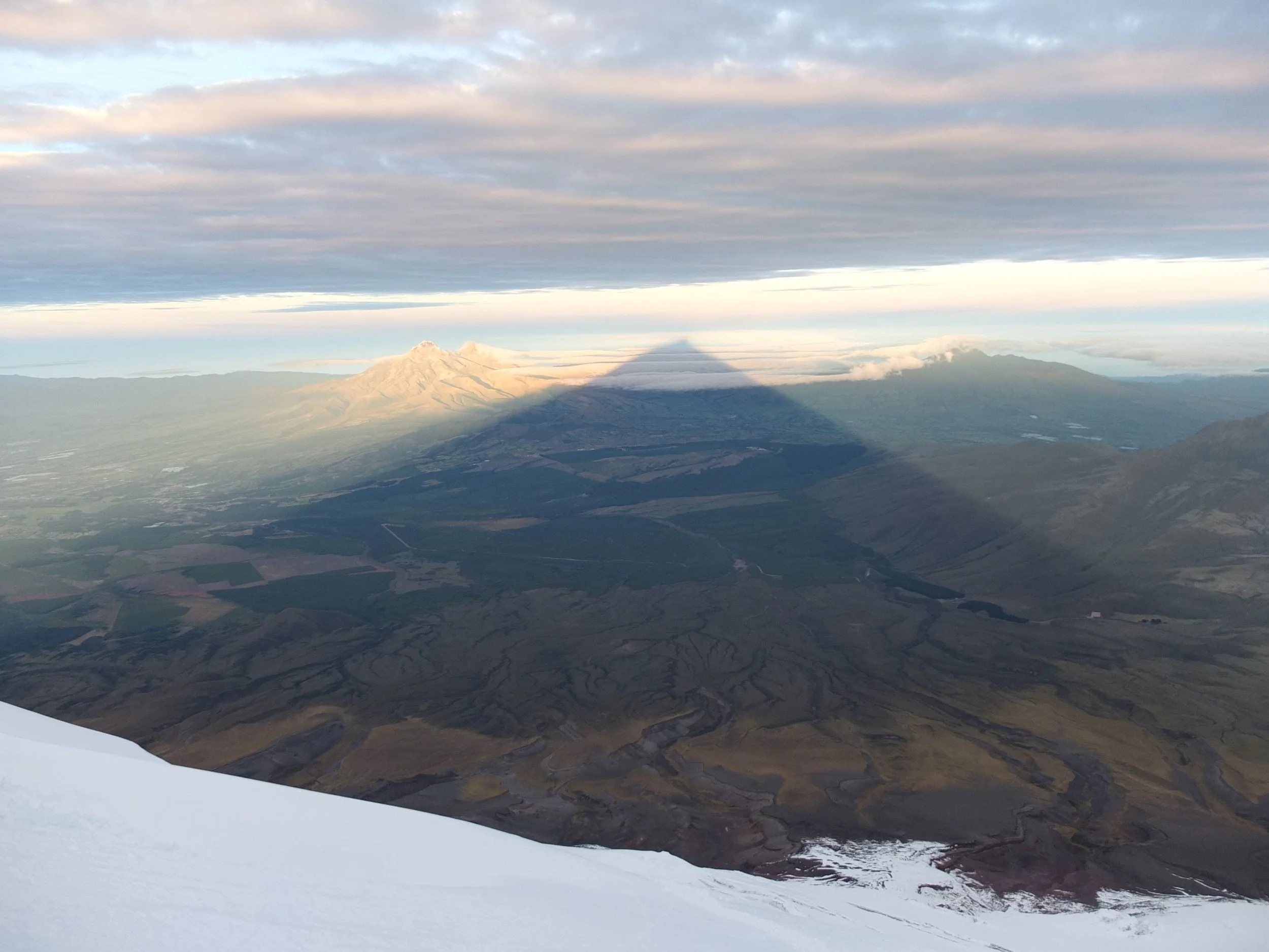 Aerial view of a snow-capped mountain with a shadow in the shape of a pyramid, surrounded by valleys, plains, and a partly cloudy sky.