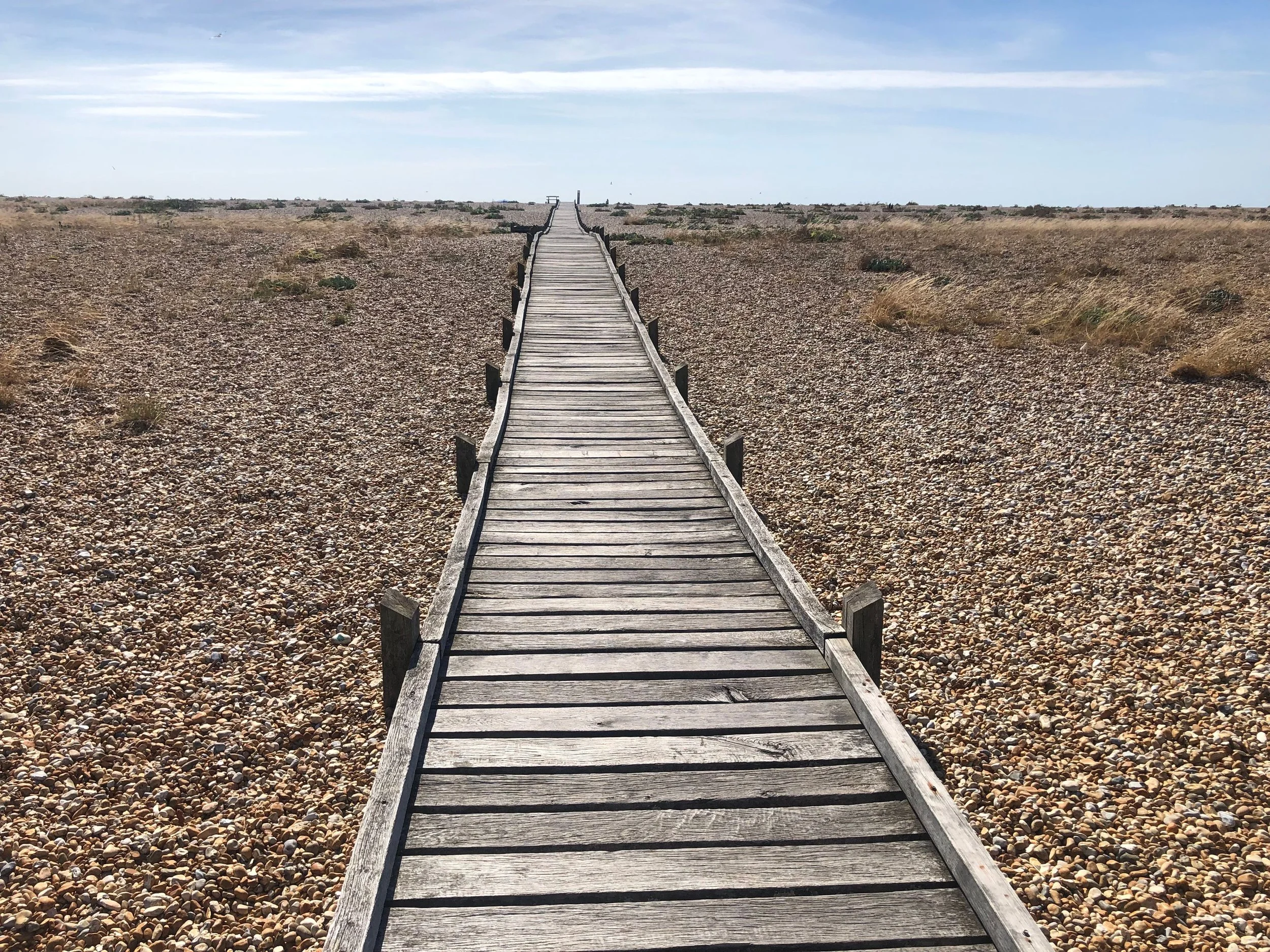 A wooden boardwalk stretches through a sandy, rocky, and sparse desert landscape under a partly cloudy sky, with distant terrain and a few small plants.