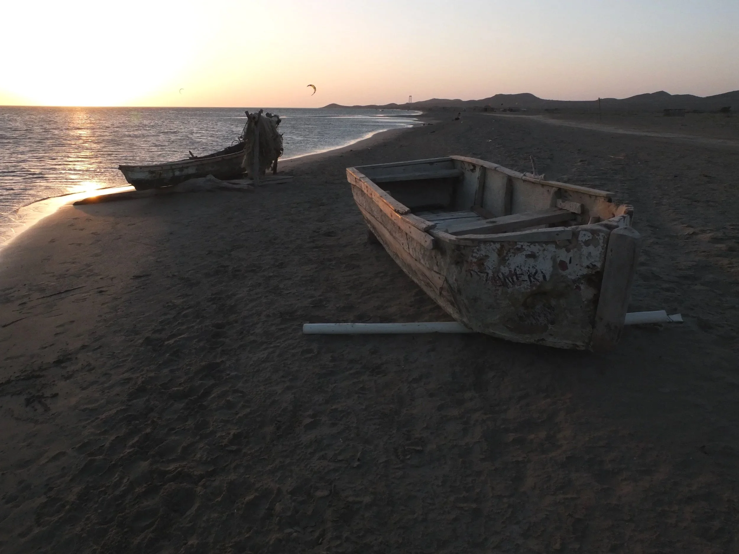 A deserted beach at sunset with two abandoned weathered boats on the sand, one upright and one overturned, along the shoreline with distant hills and kite surfers in the background.