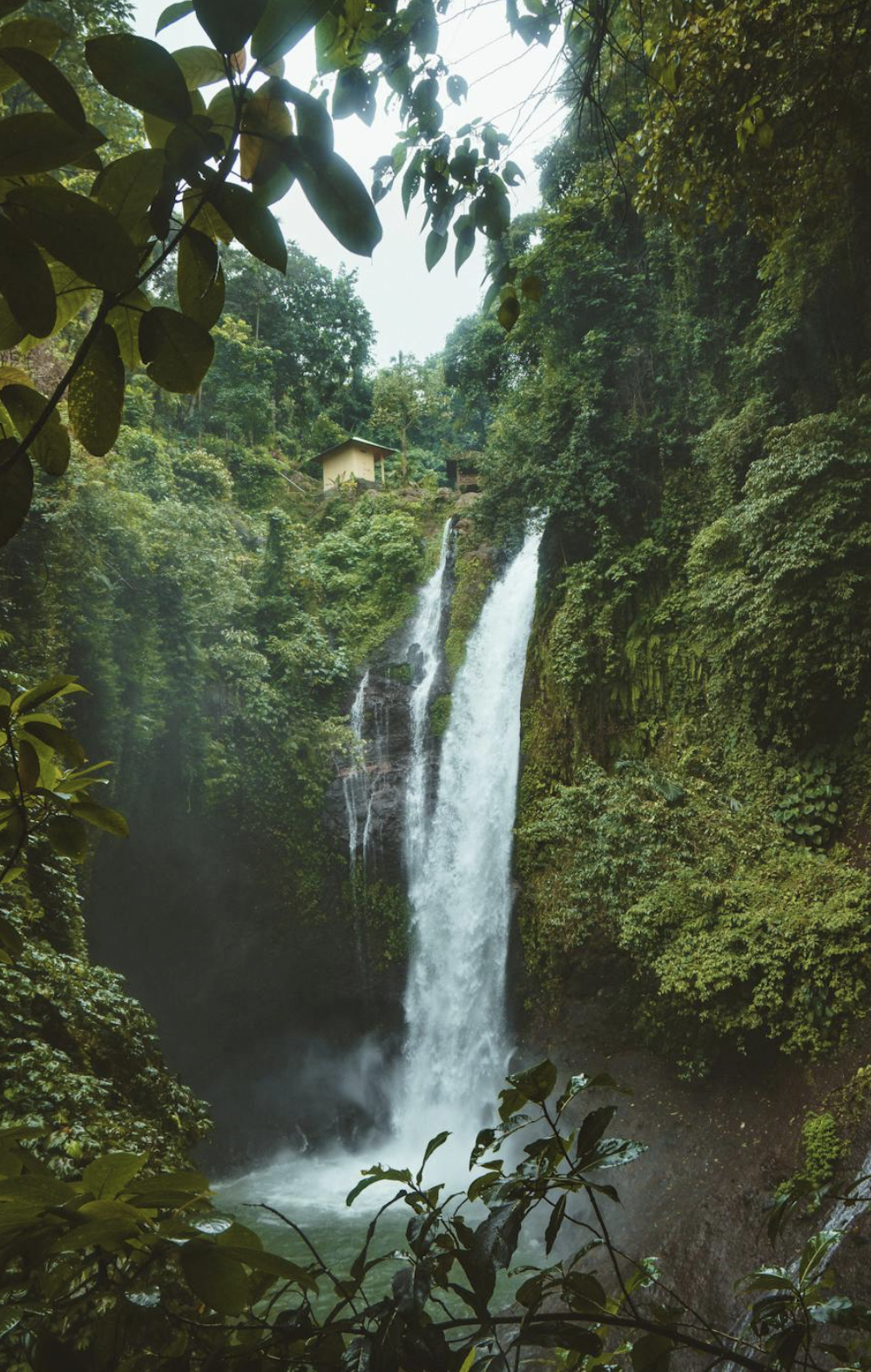 Waterfall cascading down a lush green jungle with dense foliage, a small hut on the hillside, and mist rising from the water at the bottom.