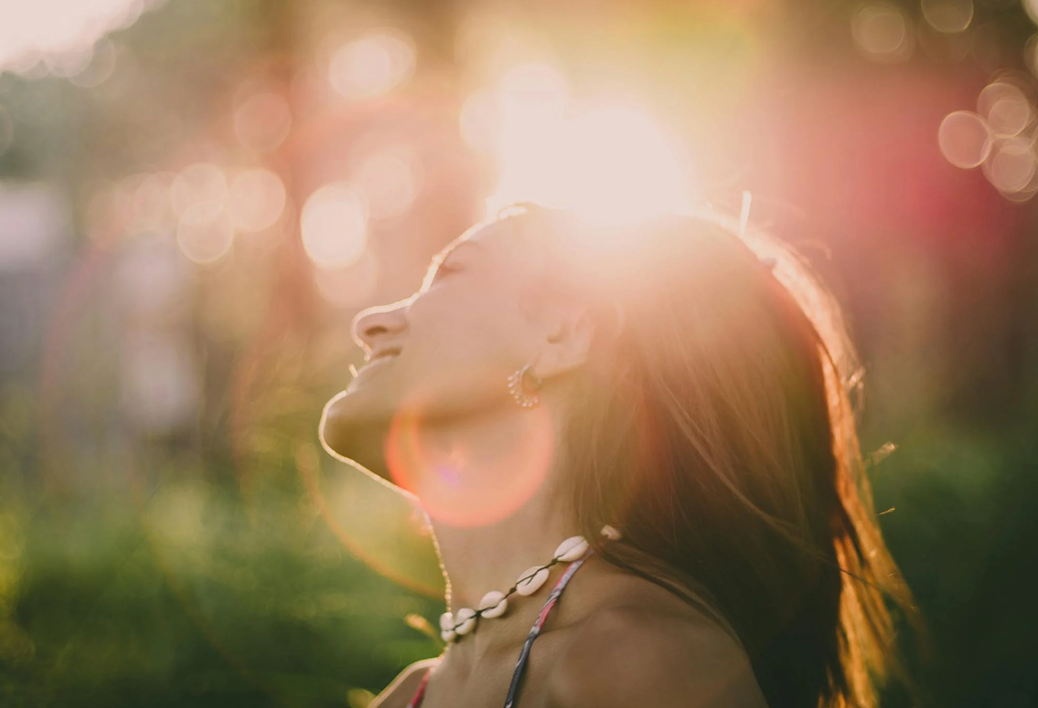 Woman with long hair wearing shell necklace and earrings, smiling with eyes closed in sunlight.