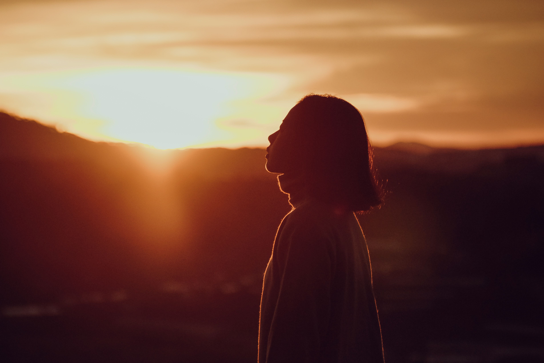 Silhouette of a girl standing outdoors during a sunset or sunrise, with the sun low on the horizon and sky filled with warm orange and yellow hues.
