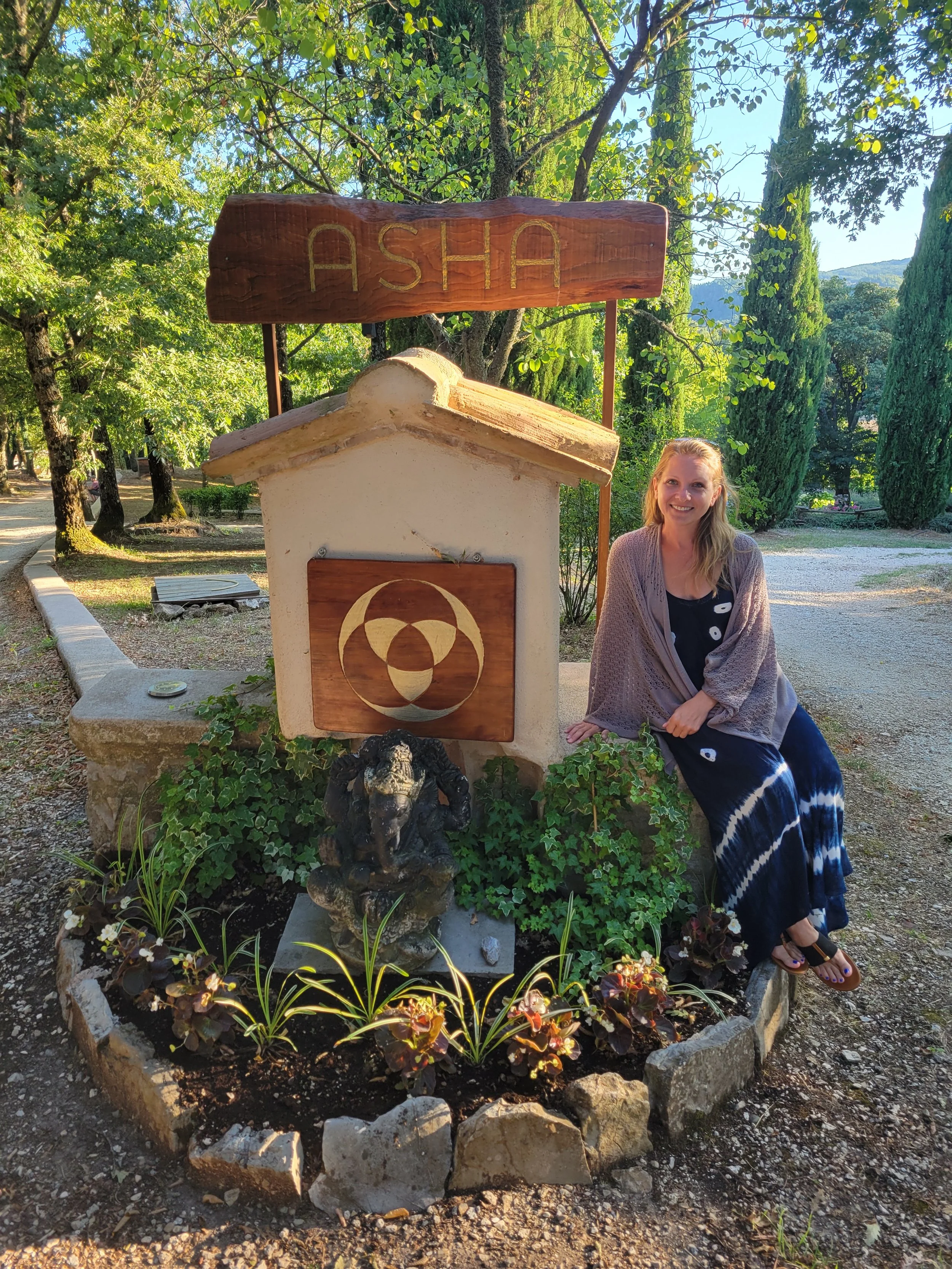 Woman sitting next to entrance sign of 'ASHA' Alchemy School of Healing Arts surrounded by trees, flowers and gravel paths.