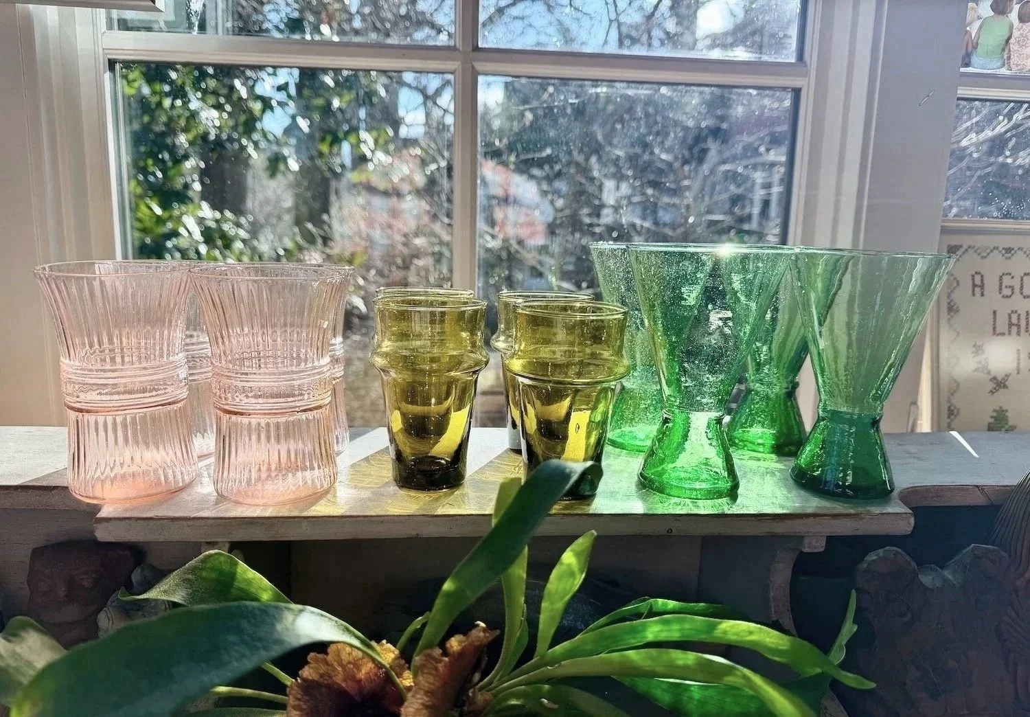 A collection of pink, yellow, and green glass cups displayed on a wooden surface in front of a window, with trees outside.