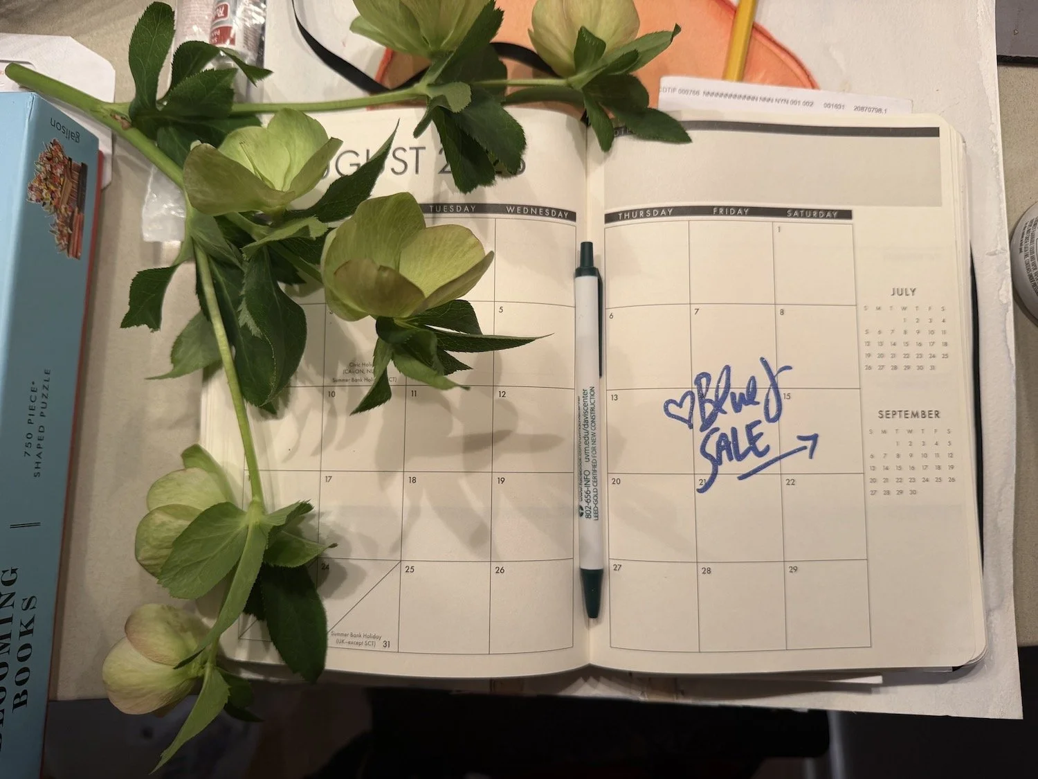 Open planner with handwritten note 'Black Sale' with hearts and arrow, accompanied by a pen. Green and white flowers draped over the planner, with surrounding objects including a box of jigsaw puzzle, a yellow pencil, and a small cylindrical container.