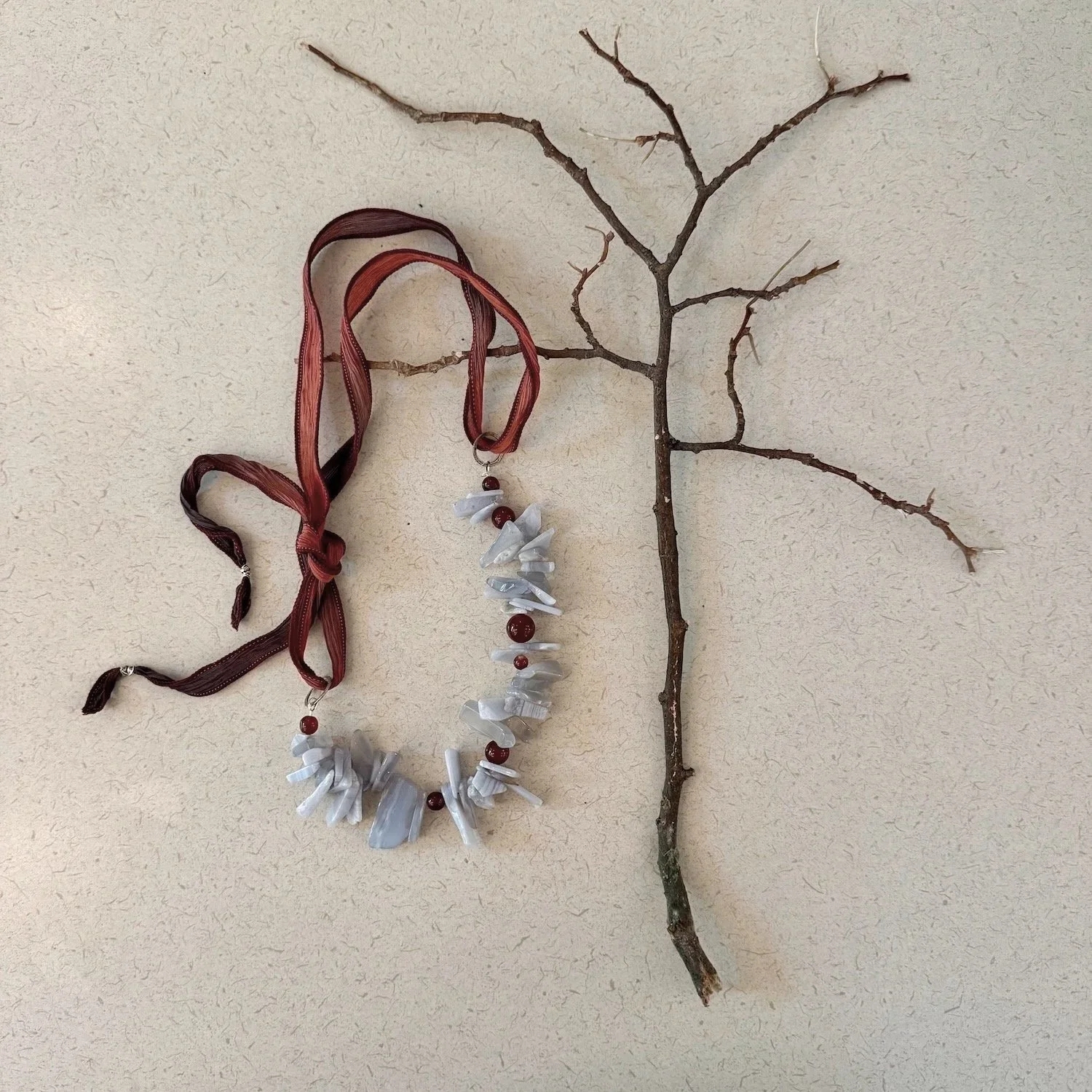 A dry, leafless tree branch mounted on a wall with two necklaces hanging from it, one with white shells and red beads, the other with light gray shell pieces and red beads.