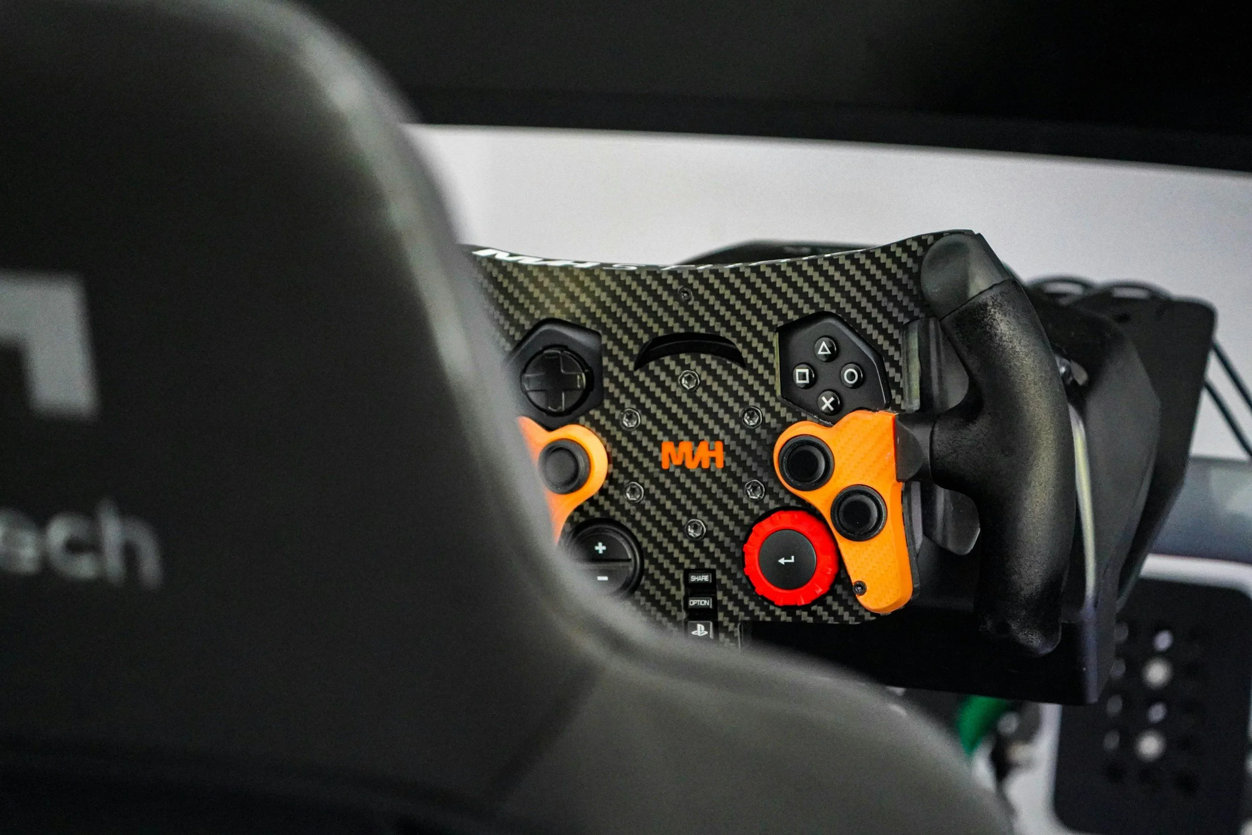Close-up of a racing car steering wheel with a black carbon fiber finish, orange buttons, and a red dial, inside a car cockpit.