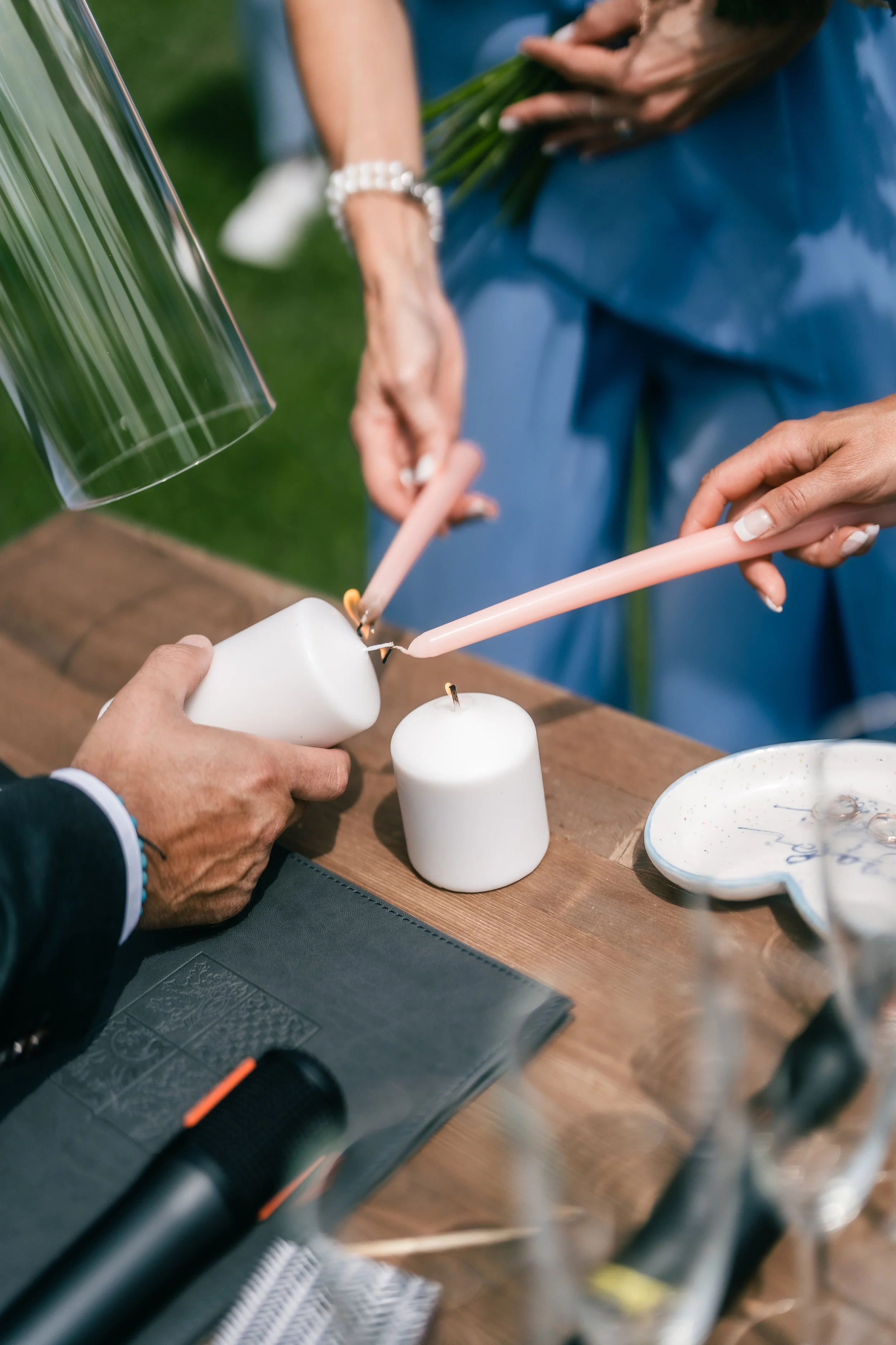 A close-up of hands lighting candles with a Bic lighter on a wooden table, featuring white candles and a decorative dish, with a person holding a bouquet of flowers in the background.