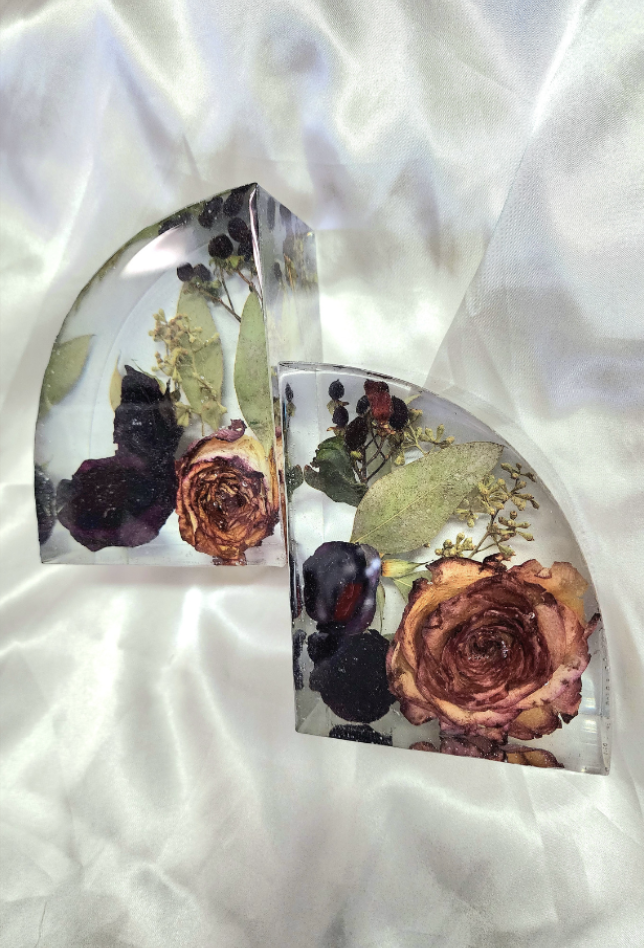 Two heart-shaped glass sculptures containing dried roses, berries, and leaves, placed on a white, silky fabric background.