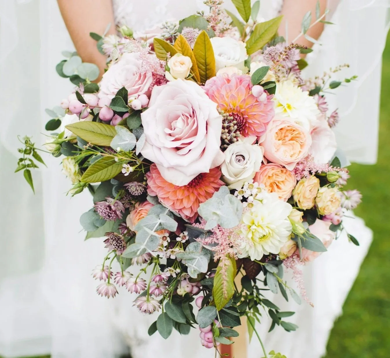 A bridal bouquet featuring pale pink, white, and peach roses, dahlias, and assorted greenery with pink berries and eucalyptus.