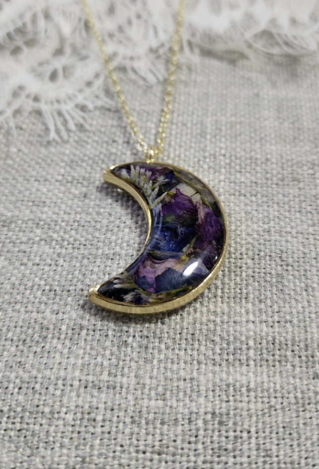 Gold crescent moon pendant with purple, green, and black pressed flowers, on a gold chain, placed on gray fabric background with lace trim.