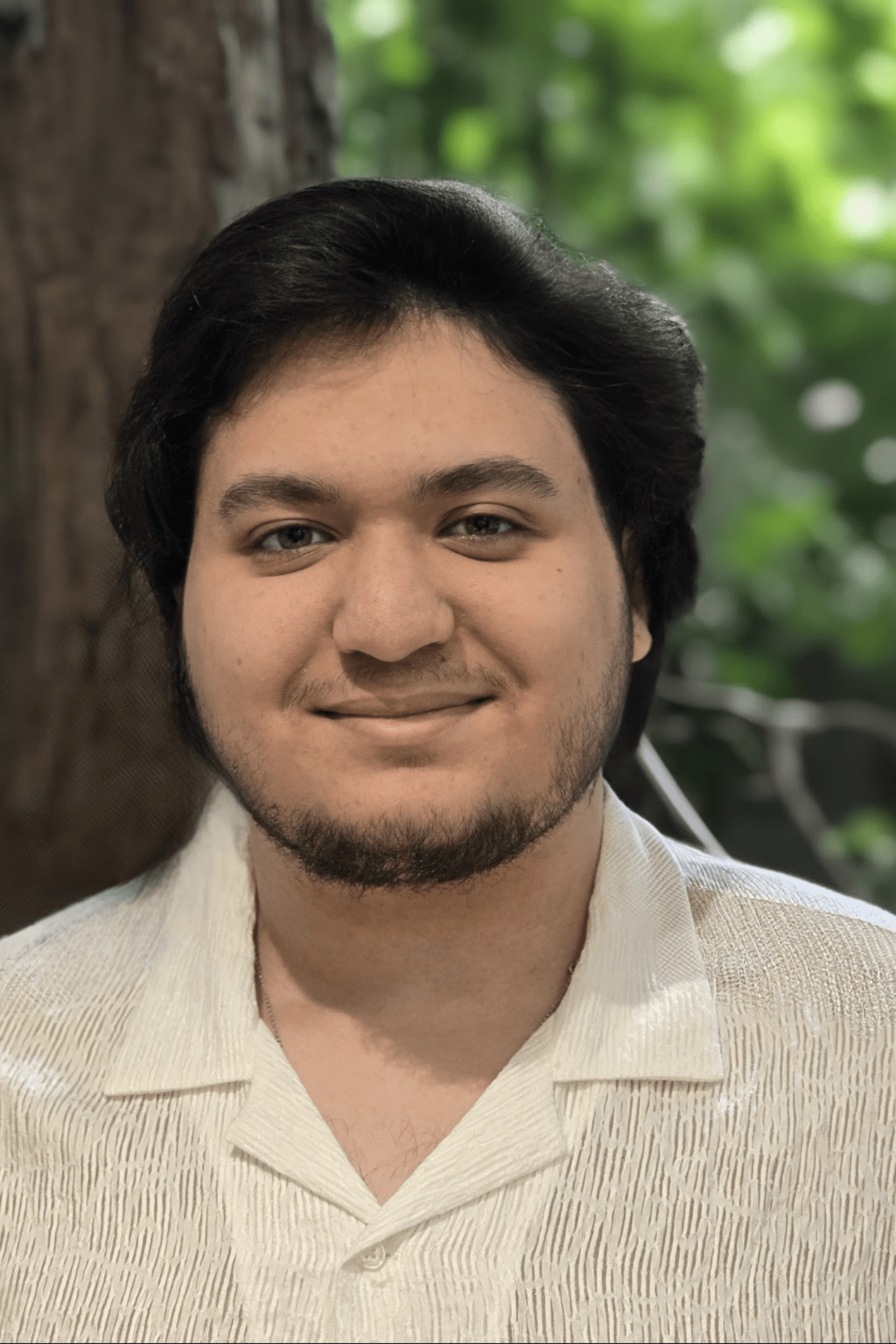 Portrait of a young man with dark hair, light beard, smiling, wearing a light-colored shirt, outdoors with a tree and green leaves in the background.