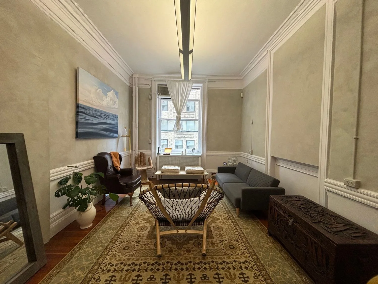 A cozy office with a large window, a gray sofa, an armchair, a wooden chest, plants, and artwork on the wall.