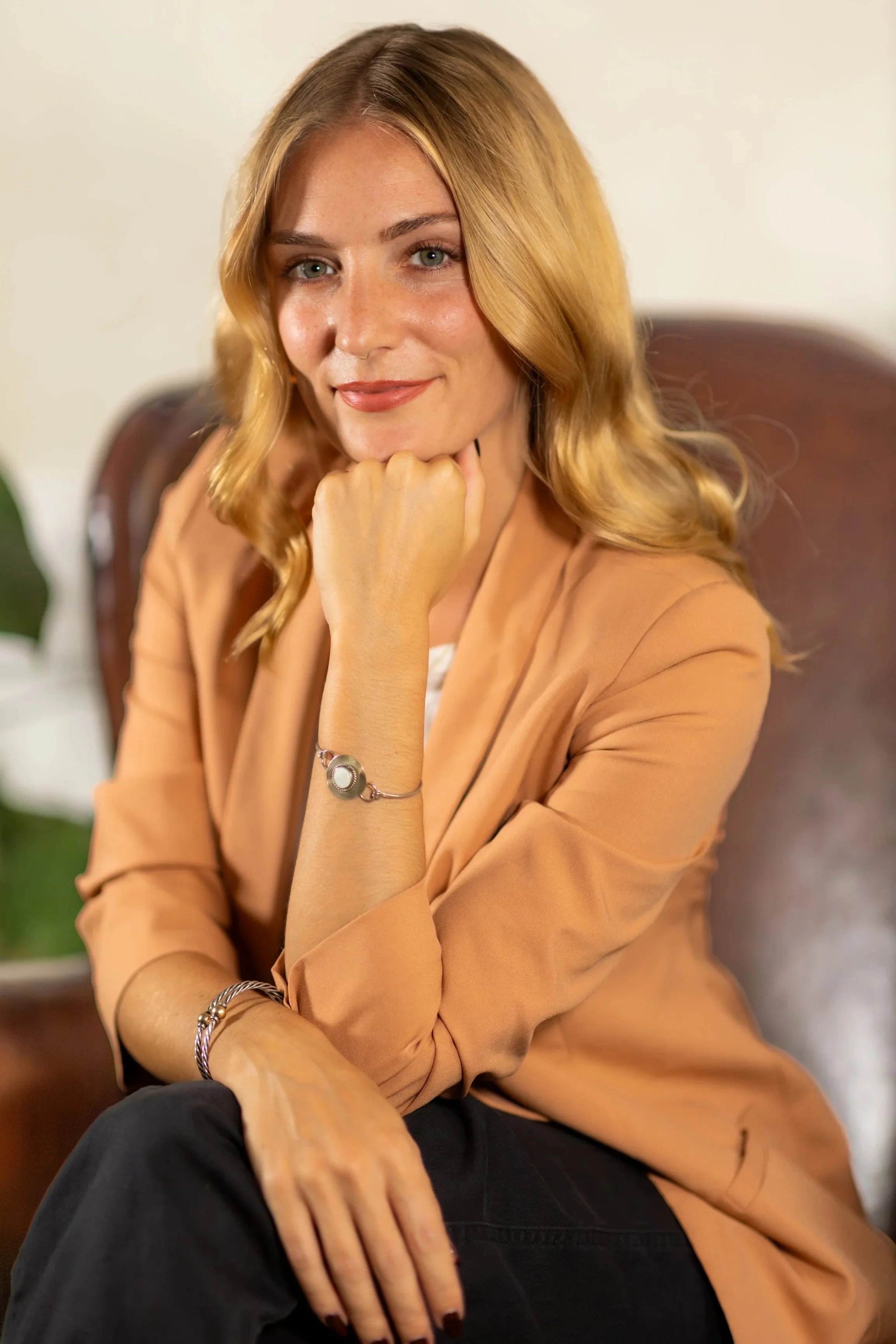 A woman with blonde hair and light skin, smiling with her chin resting on her hand, sitting on a chair indoors, wearing a beige blazer, black pants, and jewelry including a bracelet and a ring.