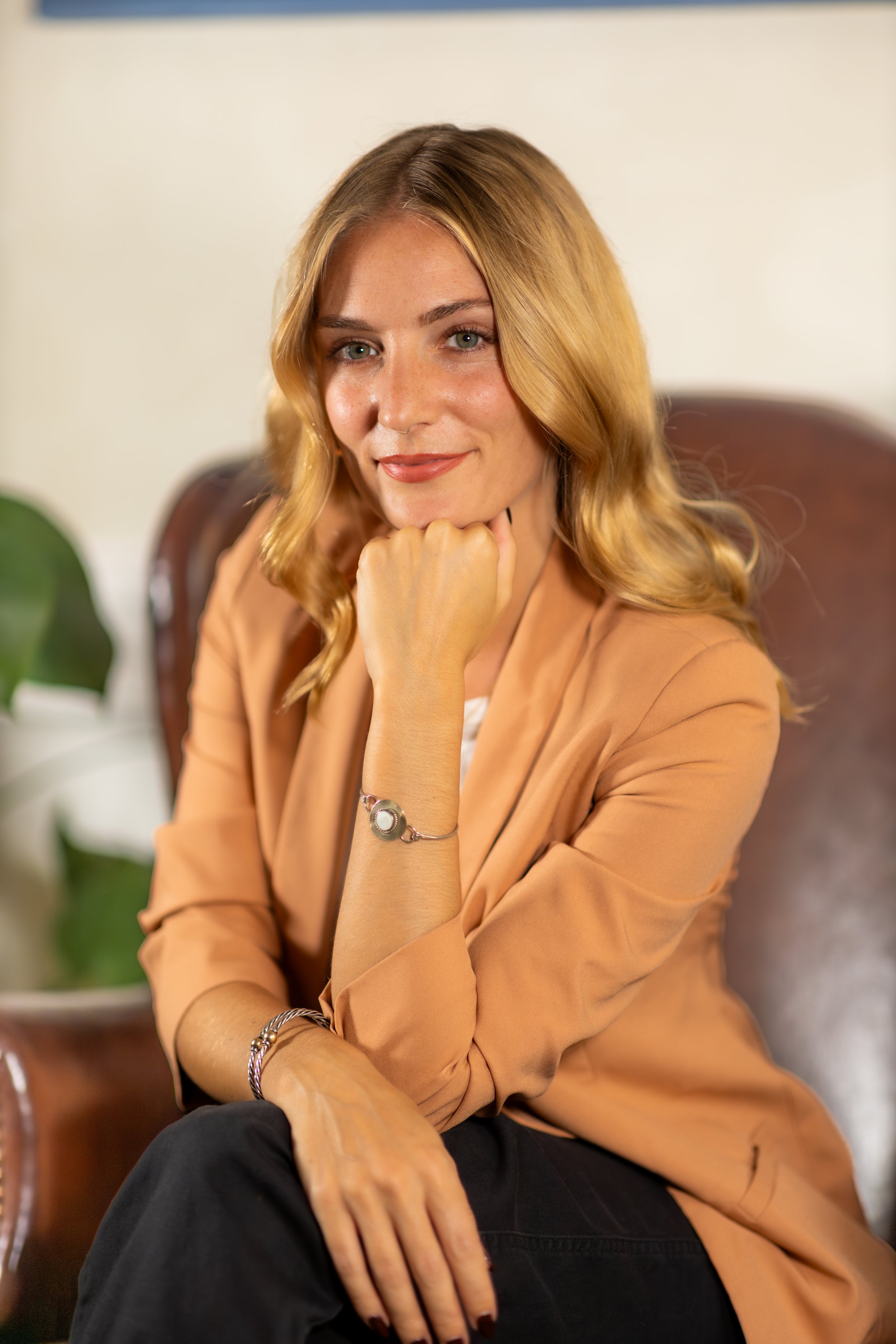 A woman with blonde hair sitting on a brown leather chair, smiling, wearing a light brown blazer, white top, and dark pants, with a silver bracelet and watch, resting her chin on her hand.