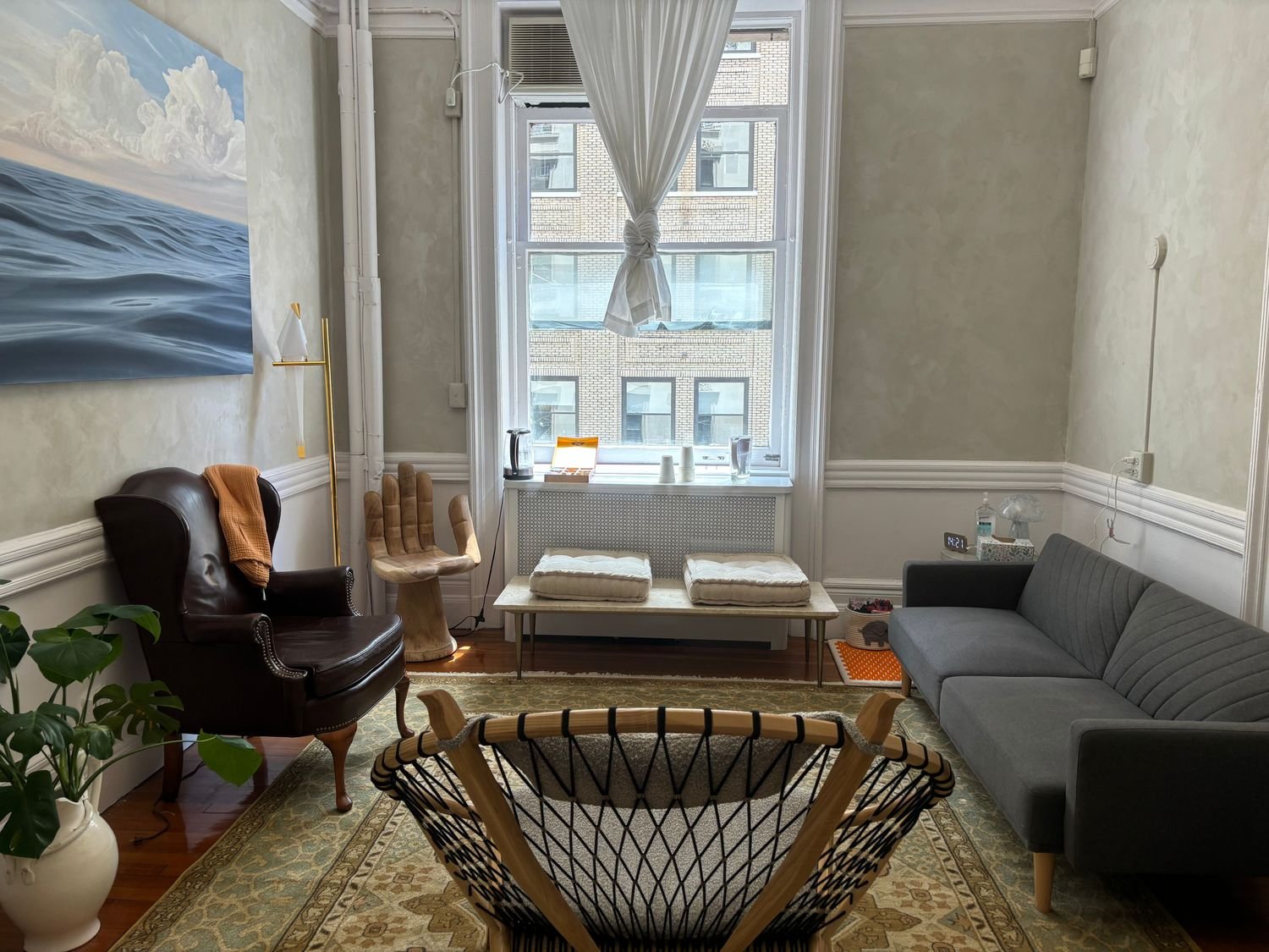 A cozy living room with a large window, a grey sofa, a black armchair with an orange throw, a wooden chair, and a woven chair. There is a painting of ocean waves on the wall, a potted plant, and various items on a table and windowsill.