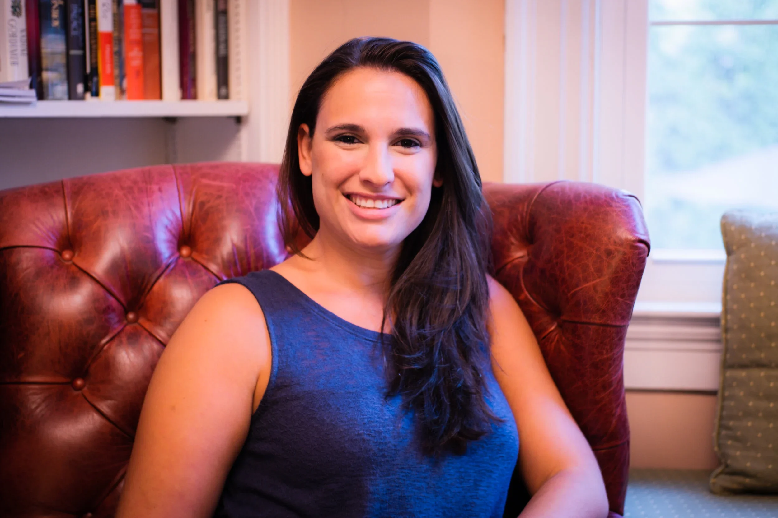 A woman with long dark hair wearing a blue sleeveless top sitting on a red leather tufted armchair in a bright room with a bookshelf and window in the background.