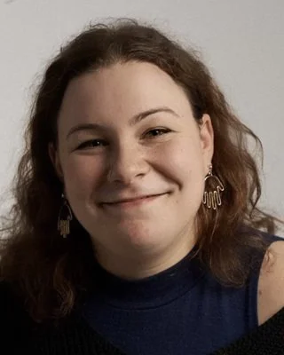 A young woman with shoulder-length brown hair smiling, wearing earrings and a dark top with a cold shoulder design.